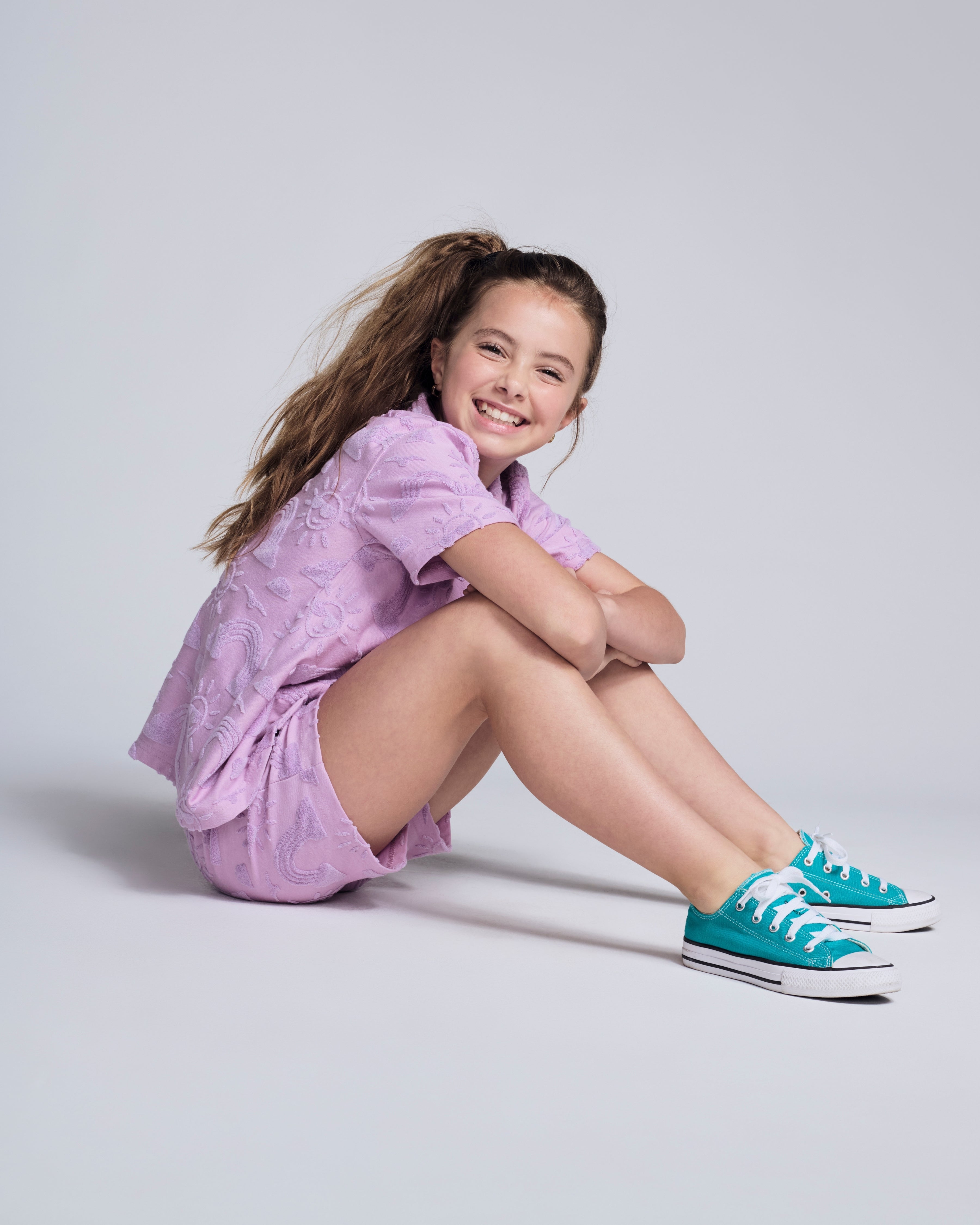 Girl wearing the Sunny Days Lilac Resort Shirt with matching shorts and teal sneakers, sitting and smiling.