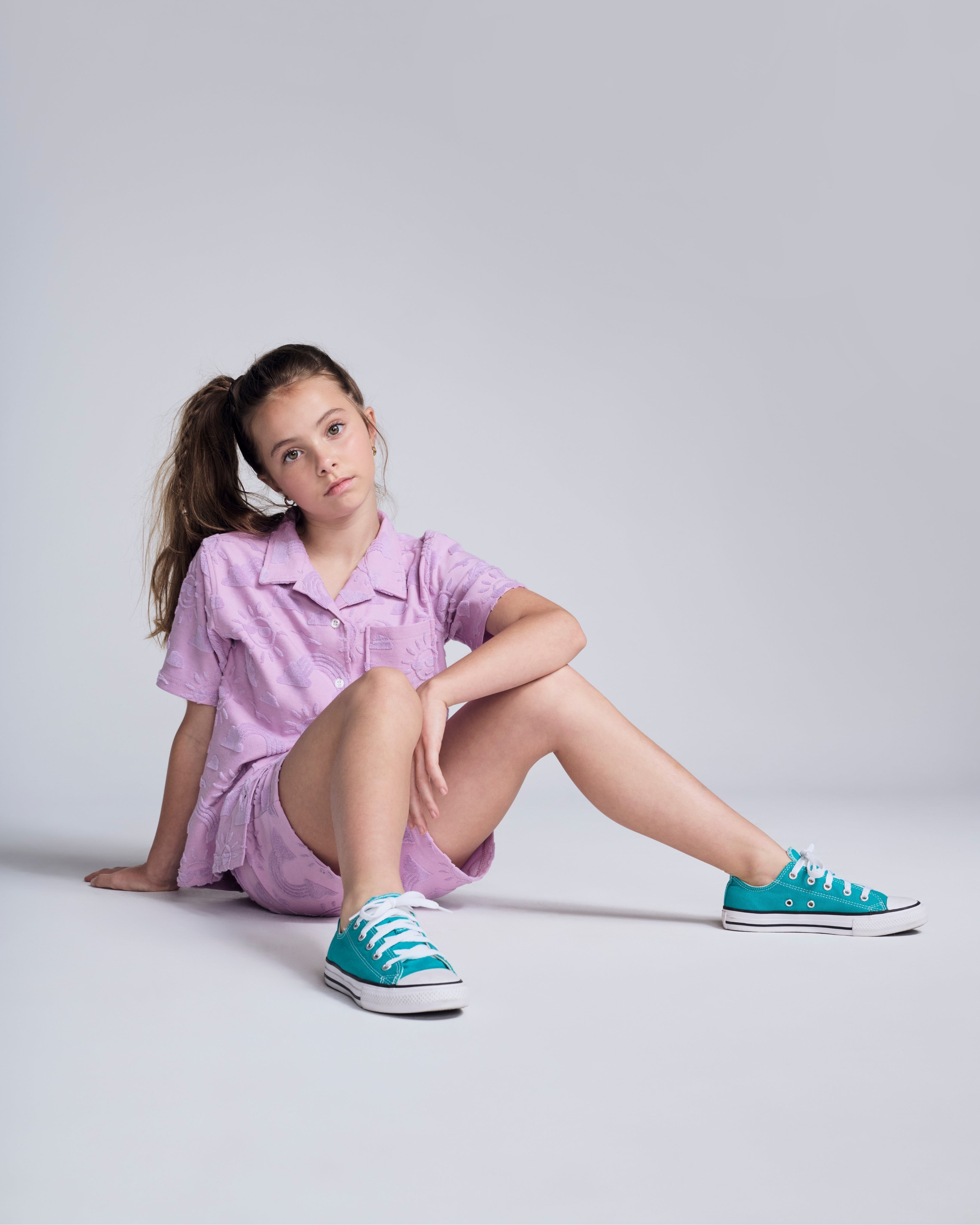 Girl wearing Sunny Days Terry Lilac Resort Short and matching shirt, styled with teal sneakers, posing in a playful manner.