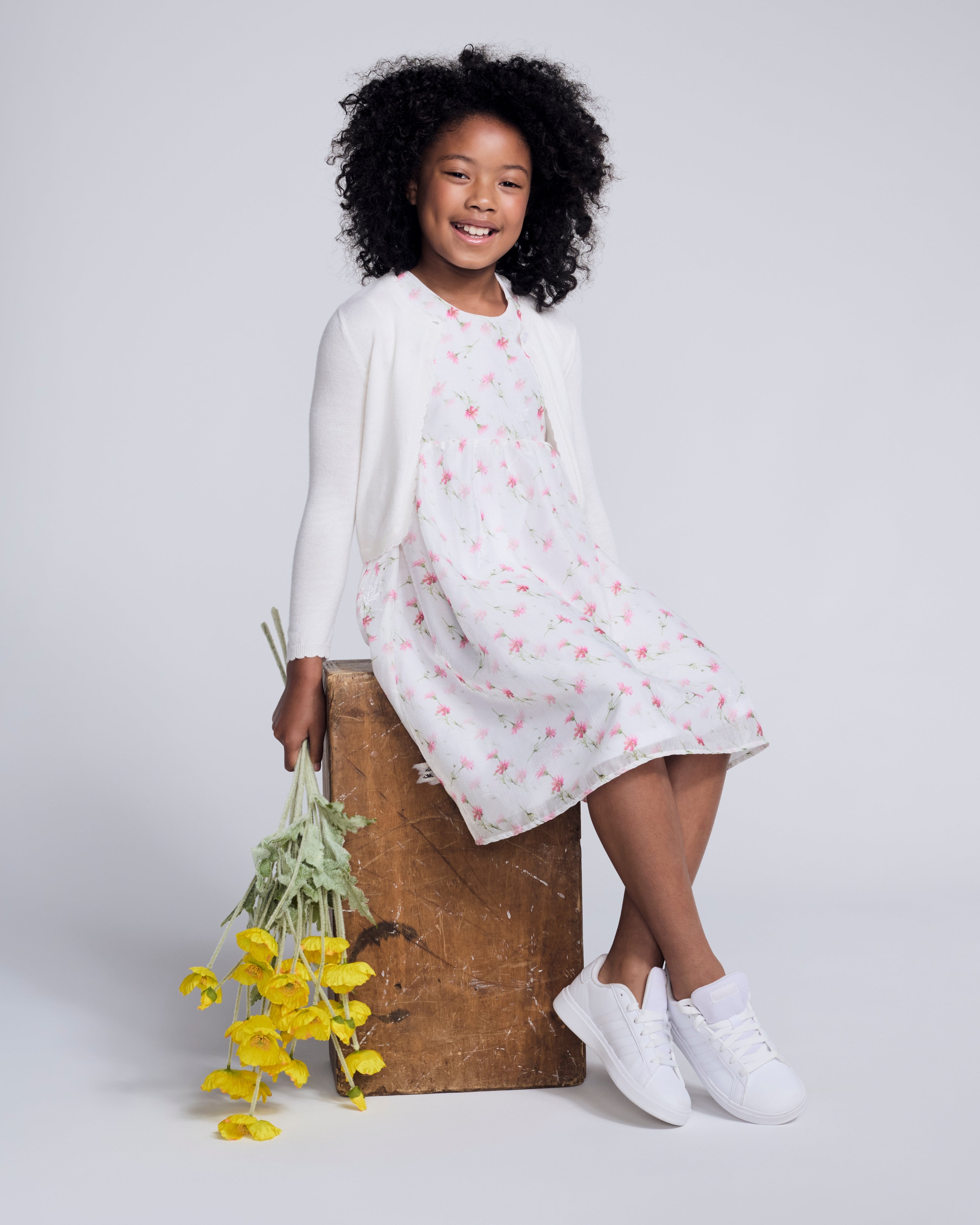 Girl wearing a White Bolero Cardigan over a floral dress, posing with flowers in hand.