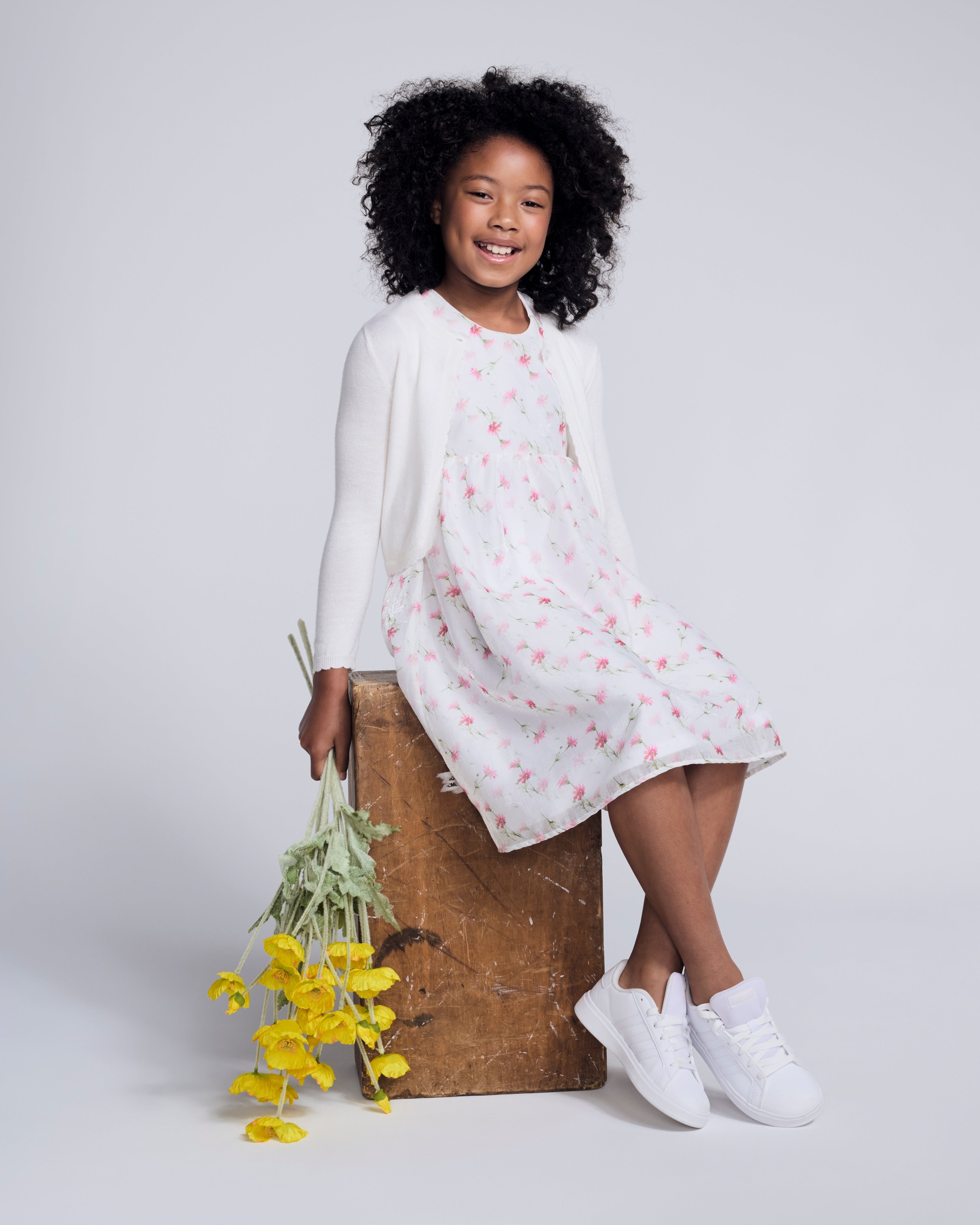 Girl wearing a White Rose Empire Dress with an allover floral print, smiling and holding flowers.