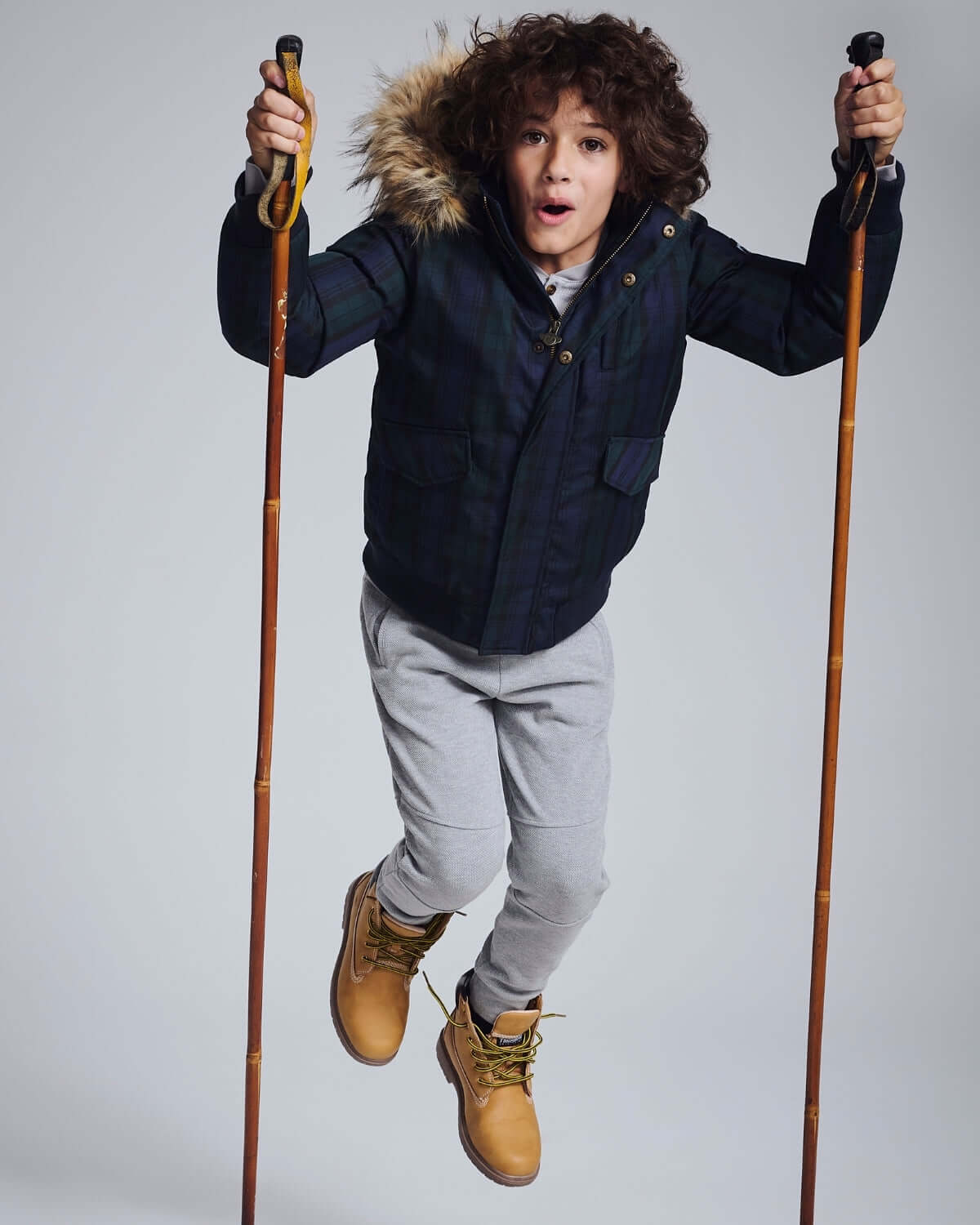 Child jumping in the Black Watch Tartan City Transit Coat with fur-lined hood and ski poles.