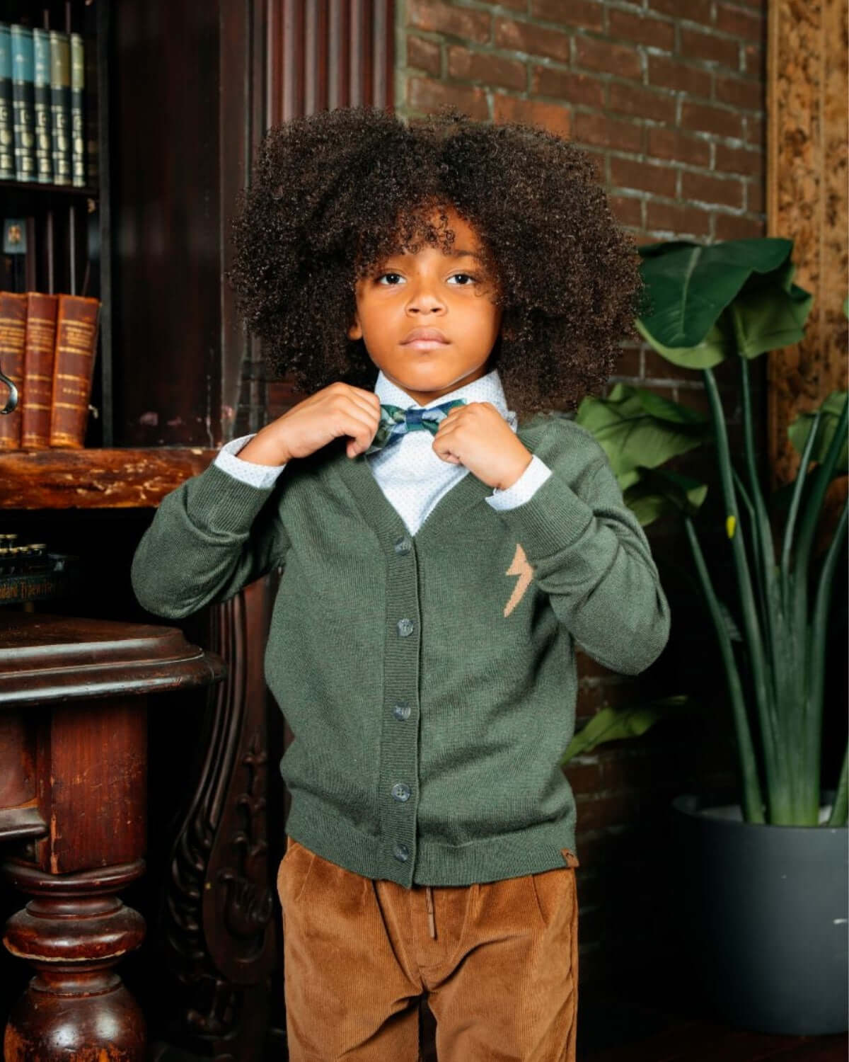Boy adjusting his bow tie, wearing British Khaki Club Pants and a stylish cardigan, showcasing elegance and comfort.
