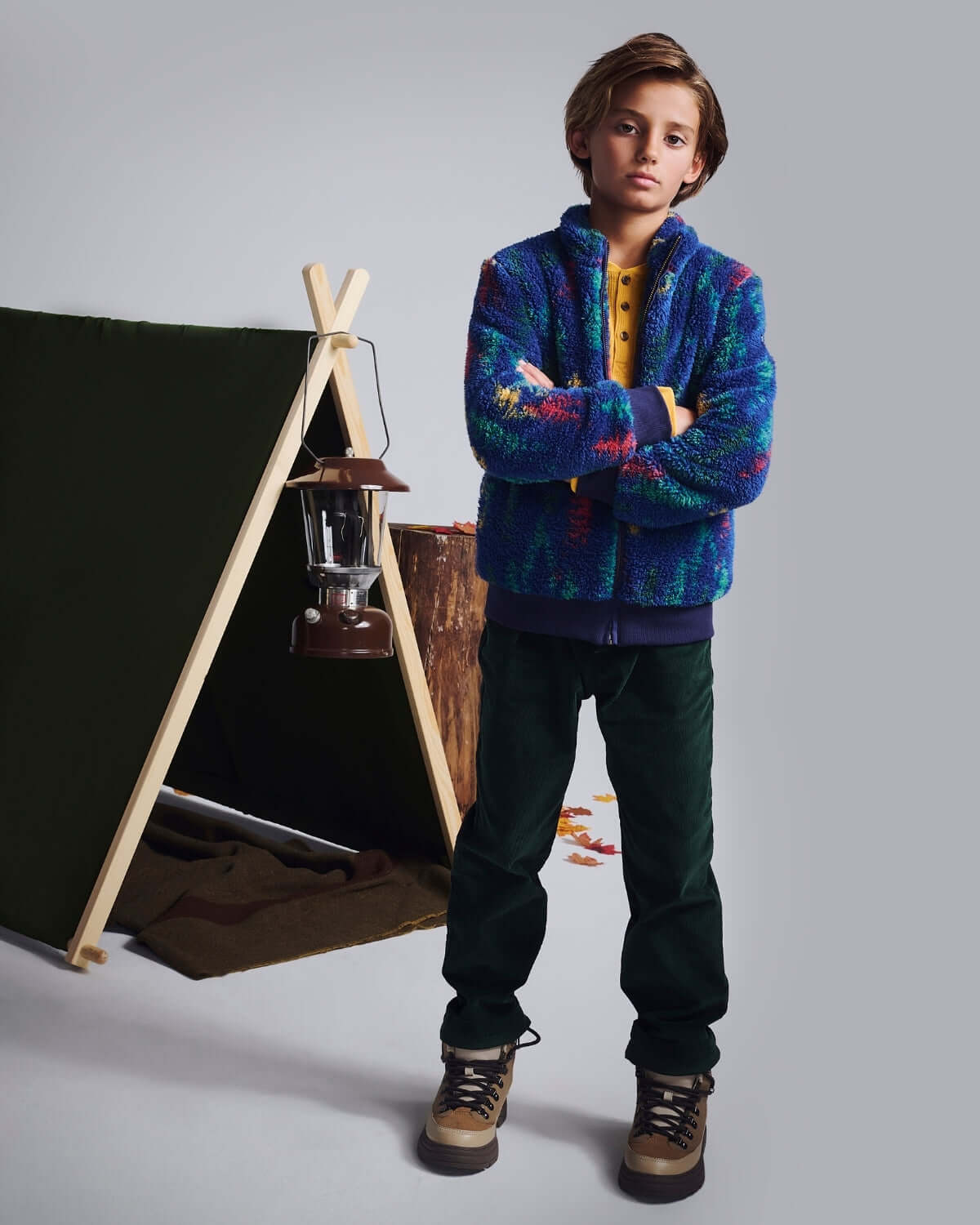 A young boy wearing Spruce Club Pants and a colorful jacket, standing in front of a tent for outdoor adventures.