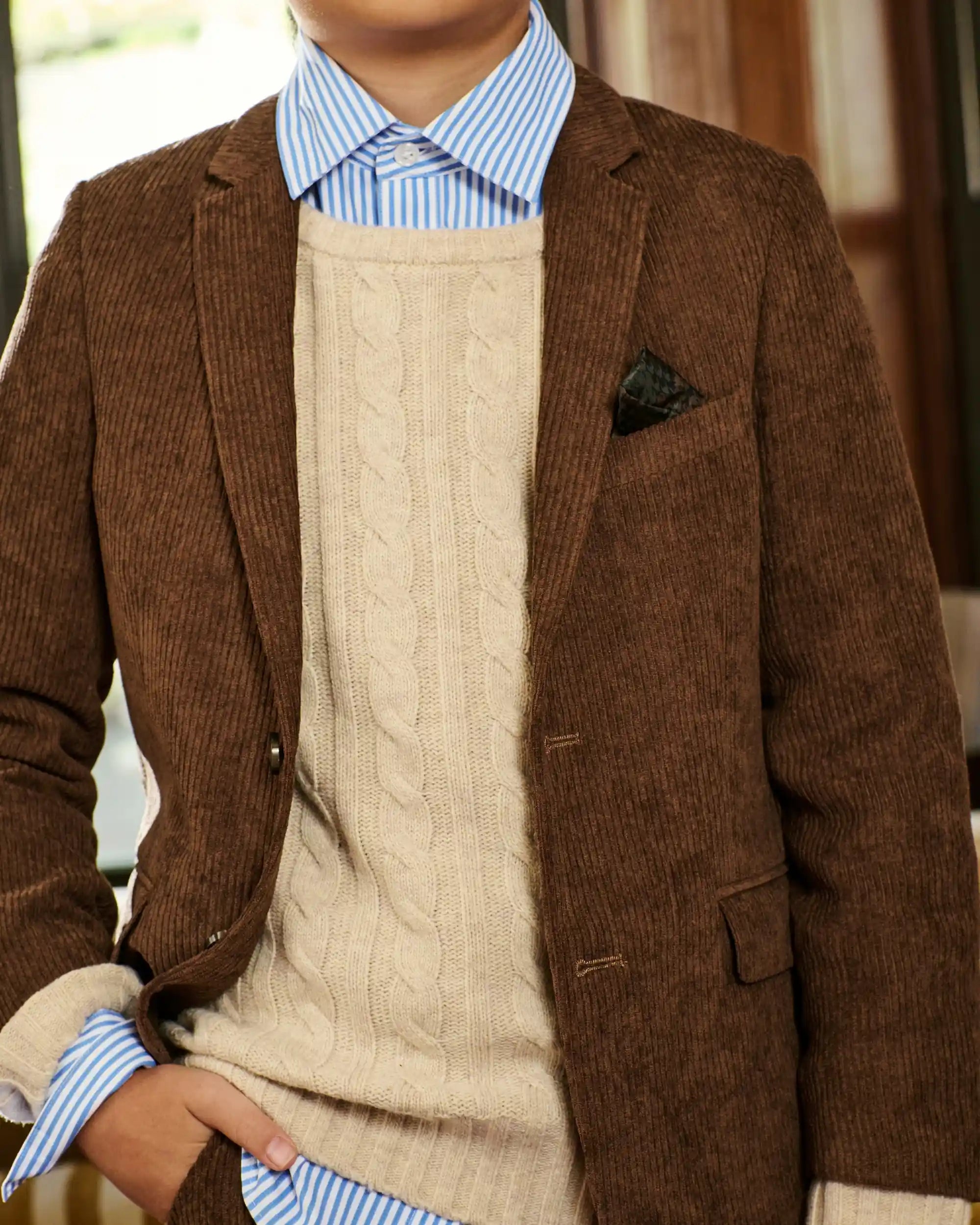 A close-up of a person wearing a brown corduroy blazer over a cream cable-knit sweater, a light blue and white striped dress shirt, and a dark pocket square.