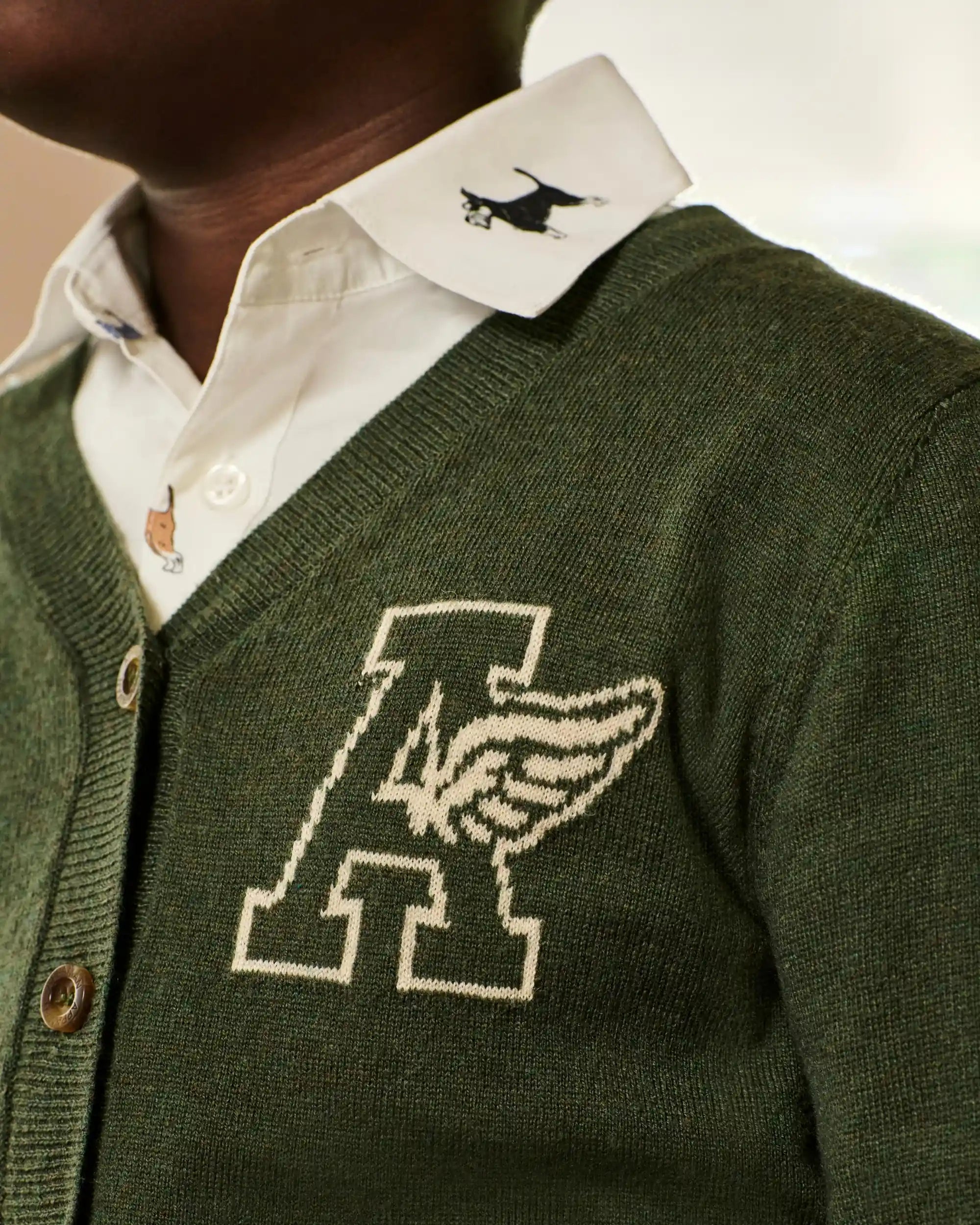Close-up of a person wearing an olive green knit cardigan over a white collared shirt. The cardigan has a large cream embroidered letter A with a wing motif on the left chest and a small black embroidered dog on the collar.