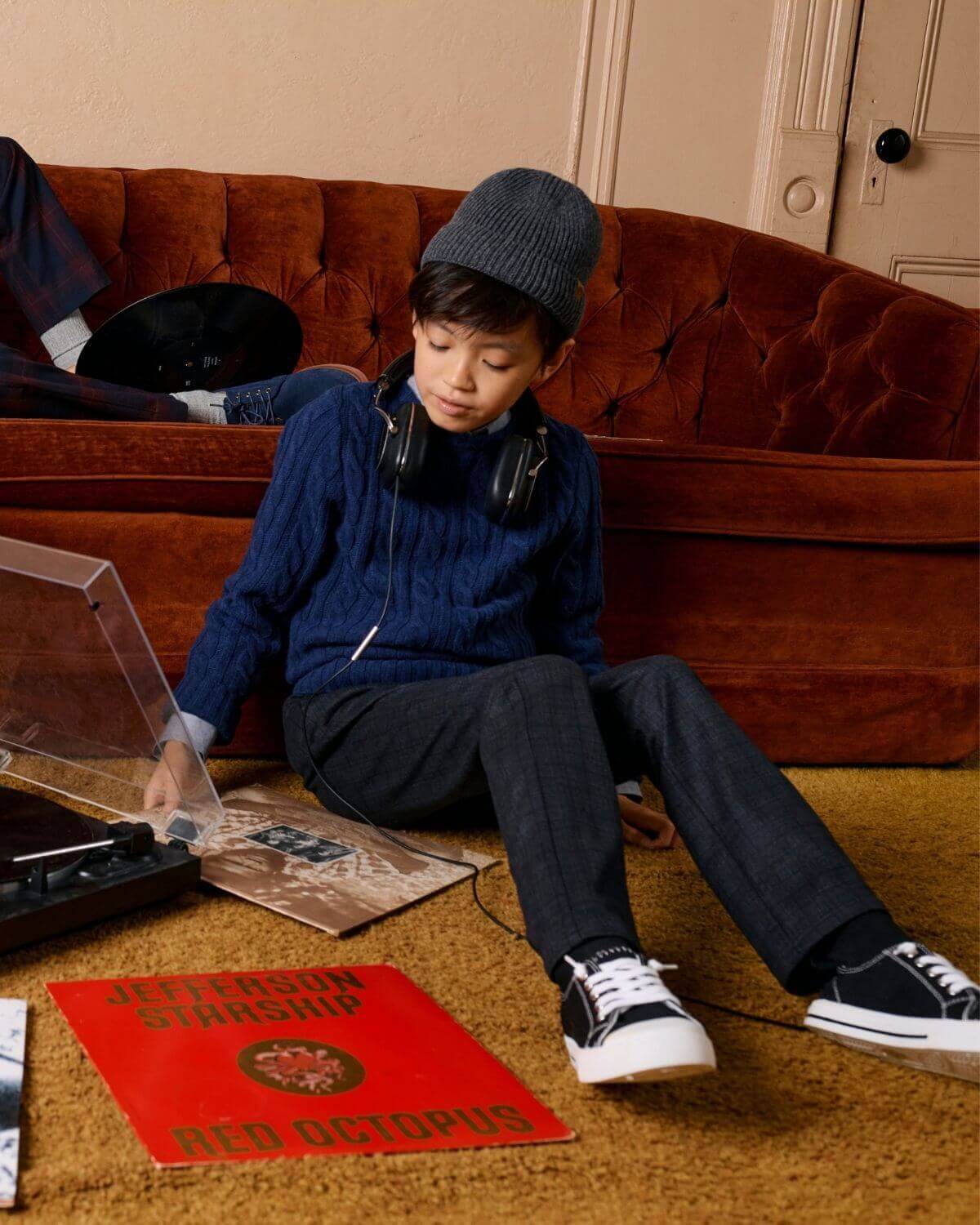 A child wearing a Navy Blue Fisherman Sweater, sitting on a couch with headphones and vinyl records.