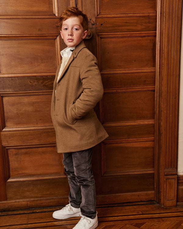 A boy wearing a camel wool coat, standing on a staircase, alternate view. Appaman Fall/Winter 2026 collection.