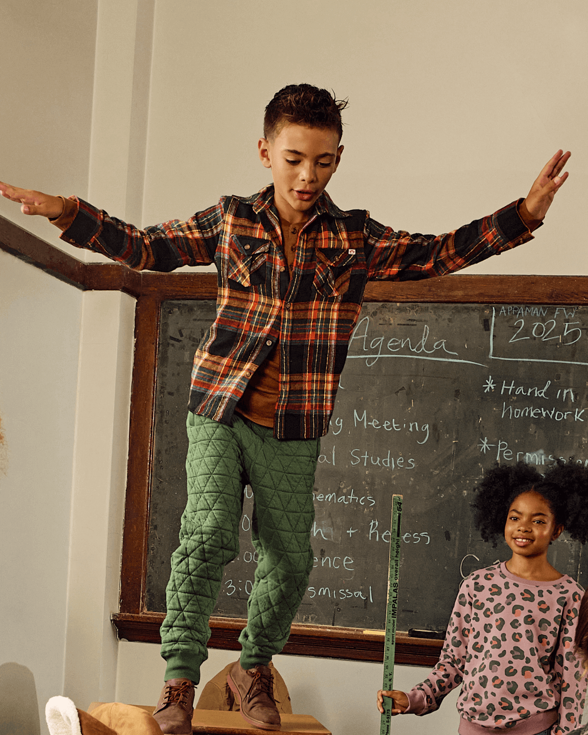 Boy balancing on a desk wearing Forest Green Highland Sweatpants and plaid shirt, showcasing cozy style in a classroom.