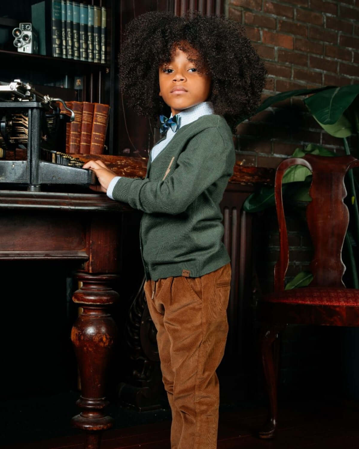 Child wearing High Club Standard Button Down shirt styled with a green cardigan, standing by a vintage desk.