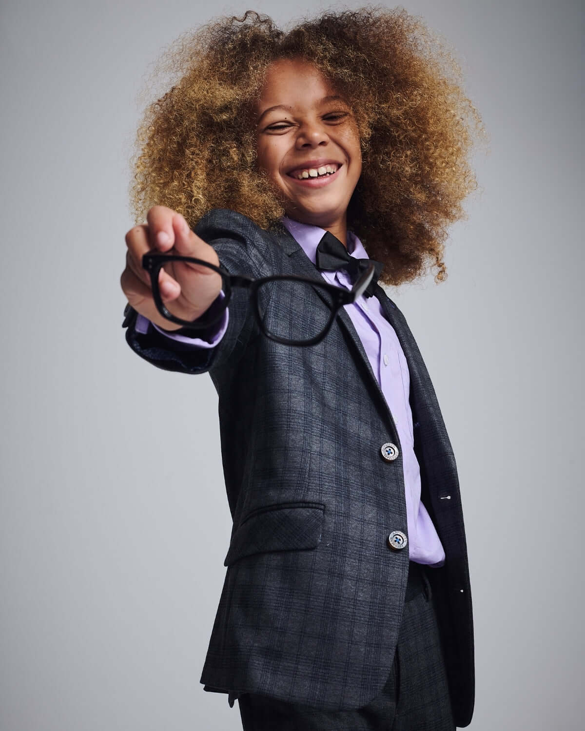 A cheerful young boy wearing a Gentleman's Plaid Stretchy Mod Suit and holding glasses, showcasing a stylish look.