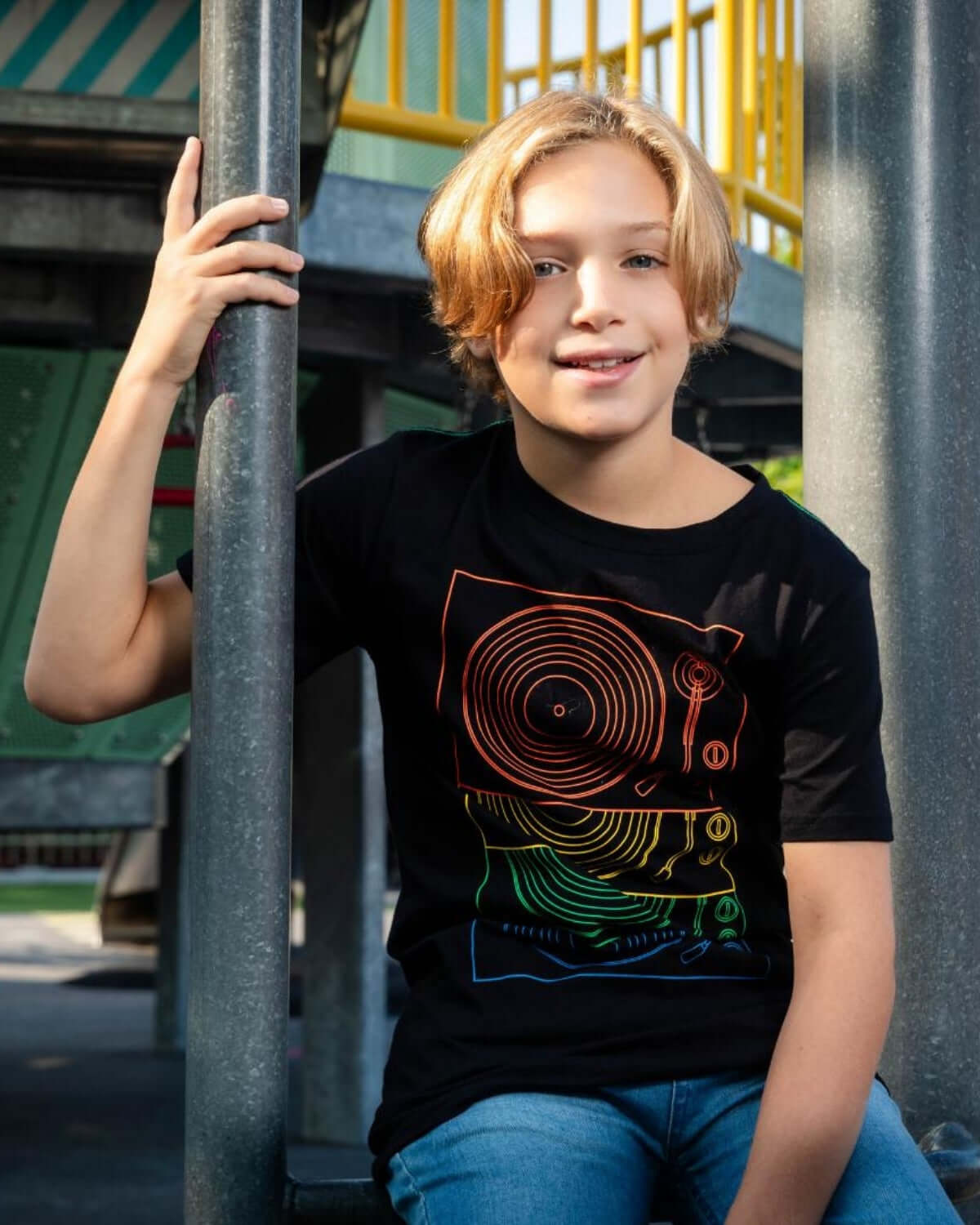 Boy wearing a Turntables Graphic Tee while sitting on a playground, showcasing vibrant DJ equipment design.