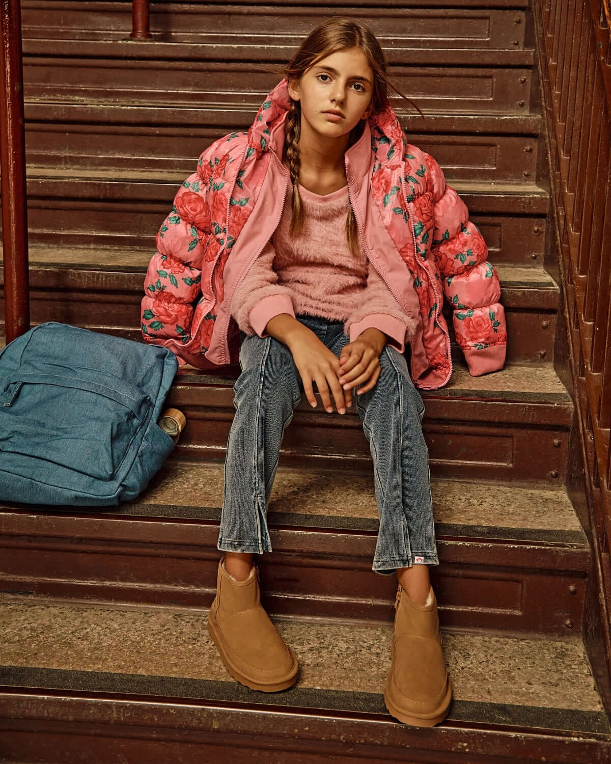 Girl sitting on stairs wearing a pink floral puffer jacket, pink sweater, denim pants, and brown boots.