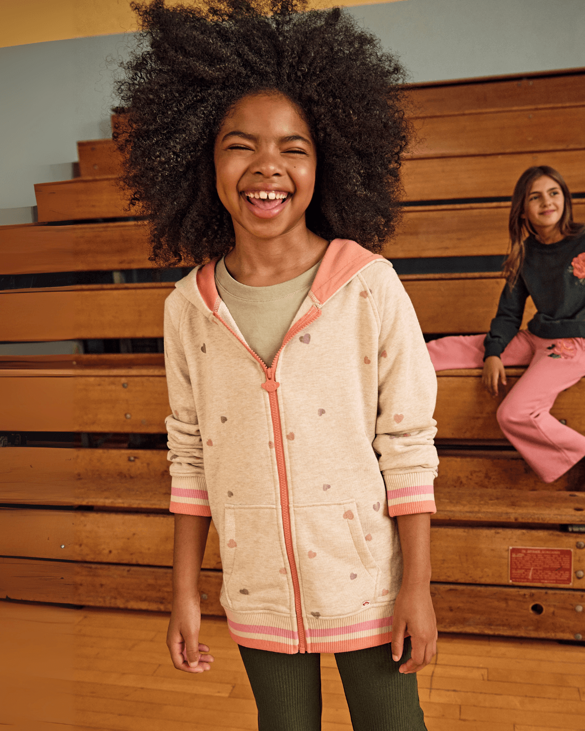 Heart Oatmeal Cozy Raglan Hoodie worn by a girl smiling in a gym setting, featuring pink and orange heart patterns.
