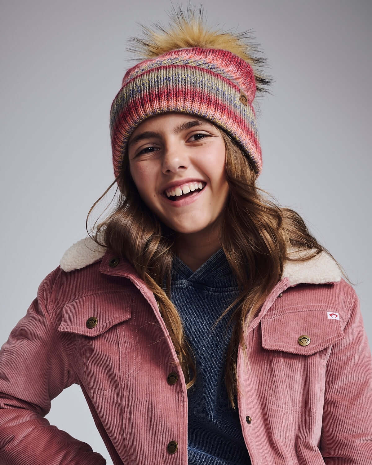 Happy girl wearing a Rose Iris Pom Beanie with a playful pom pom, styled in pink, red, and beige tones.