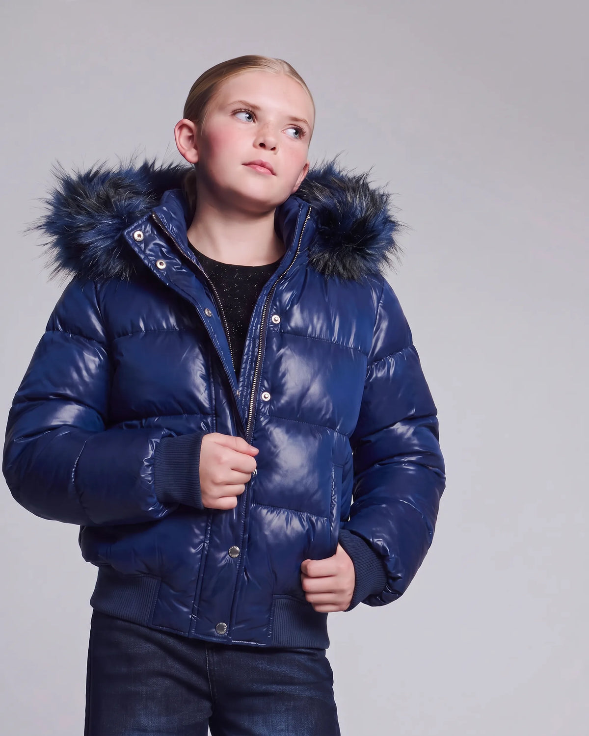 A child wearing a shiny navy blue puffer jacket with a dark faux-fur hood, photographed against a plain light gray studio background.