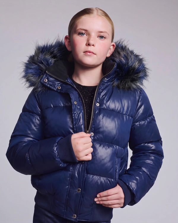 A young girl wearing a shiny navy blue puffer jacket with a dark faux-fur trimmed hood, standing against a light gray studio background.