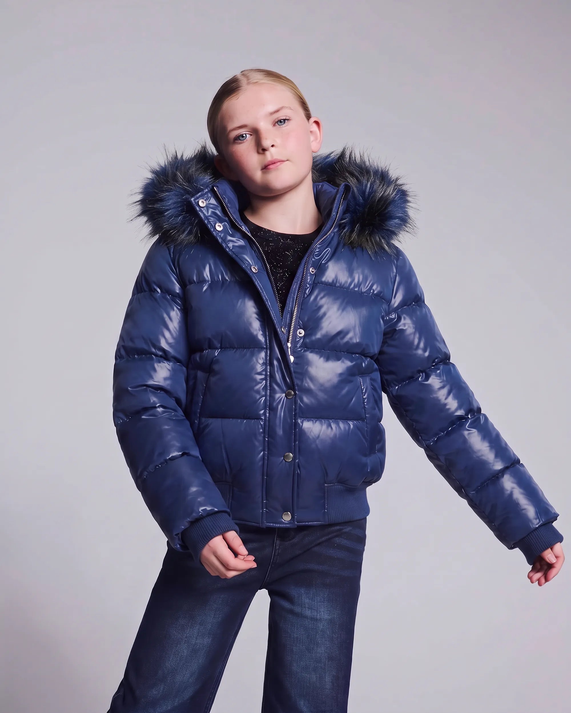 A young girl modeling a shiny navy blue puffer jacket with a dark faux-fur trimmed hood, paired with dark blue jeans, against a plain light gray studio background.