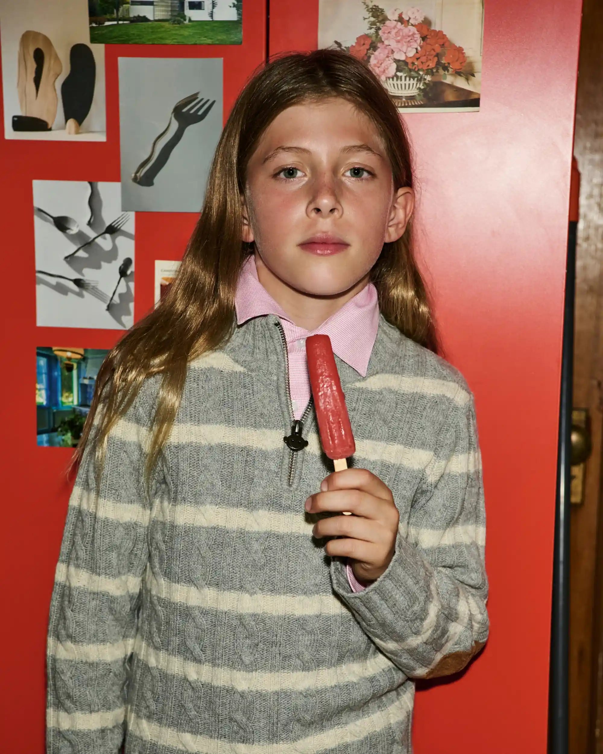 A young girl with long light brown hair stands in front of a red wall, holding a red popsicle. She is wearing a gray and white striped sweater over a pink collared shirt.