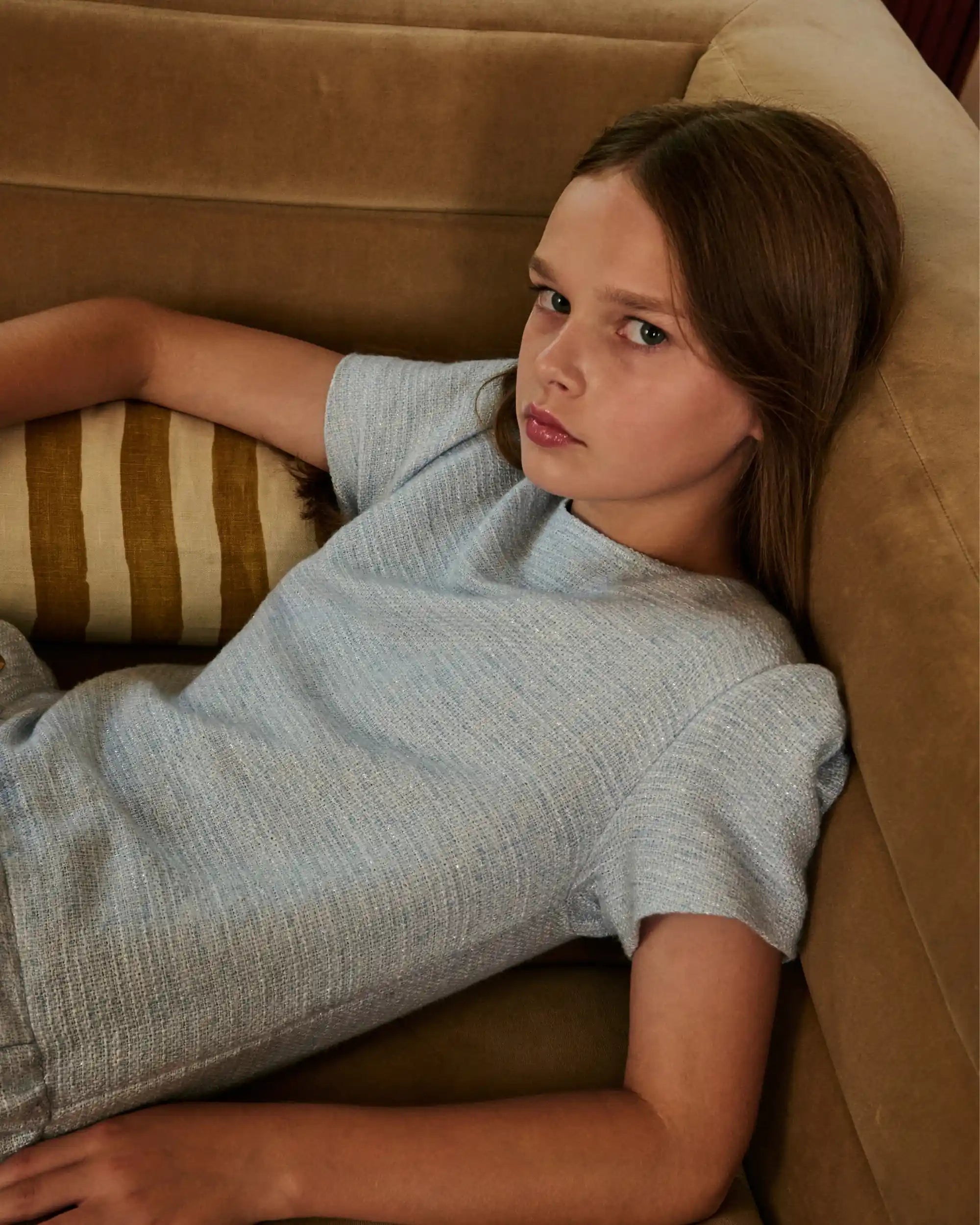 A young girl with light brown hair lies on a brown velvet sofa, wearing a light gray heathered short-sleeve t-shirt and matching shorts.