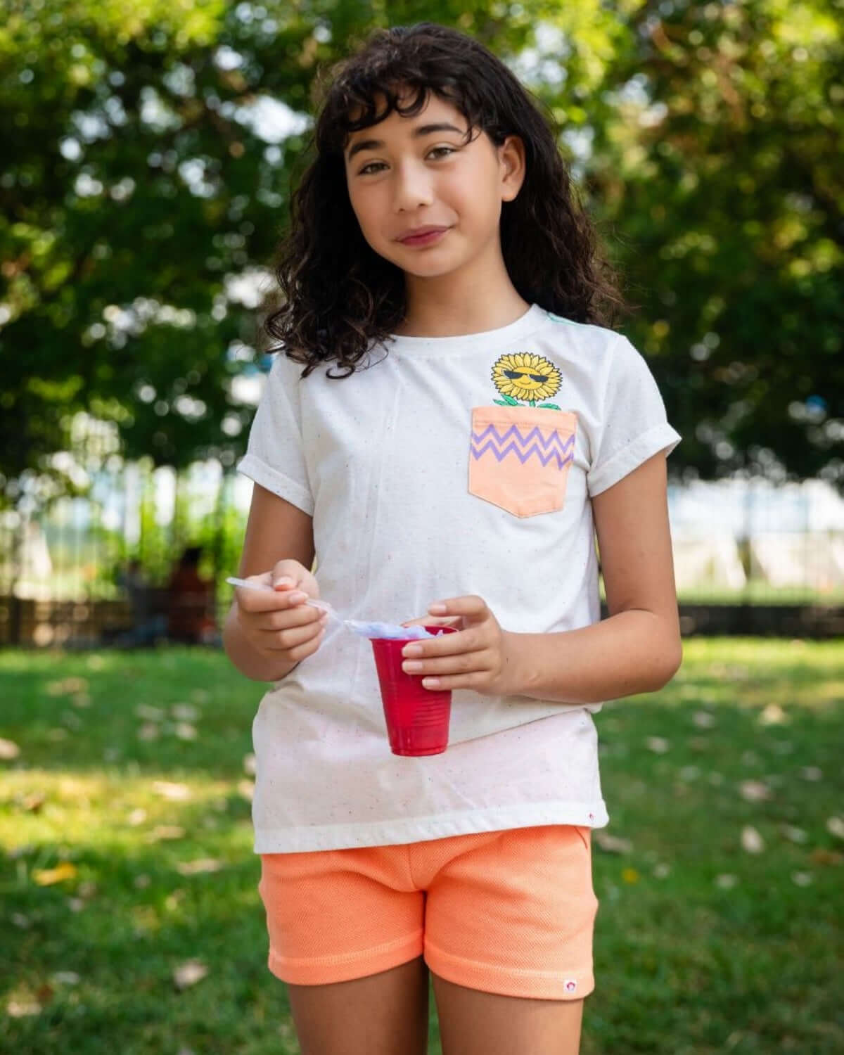 Girl wearing Peach Majorca Shorts and a light shirt, enjoying a drink outdoors in a sunny park.