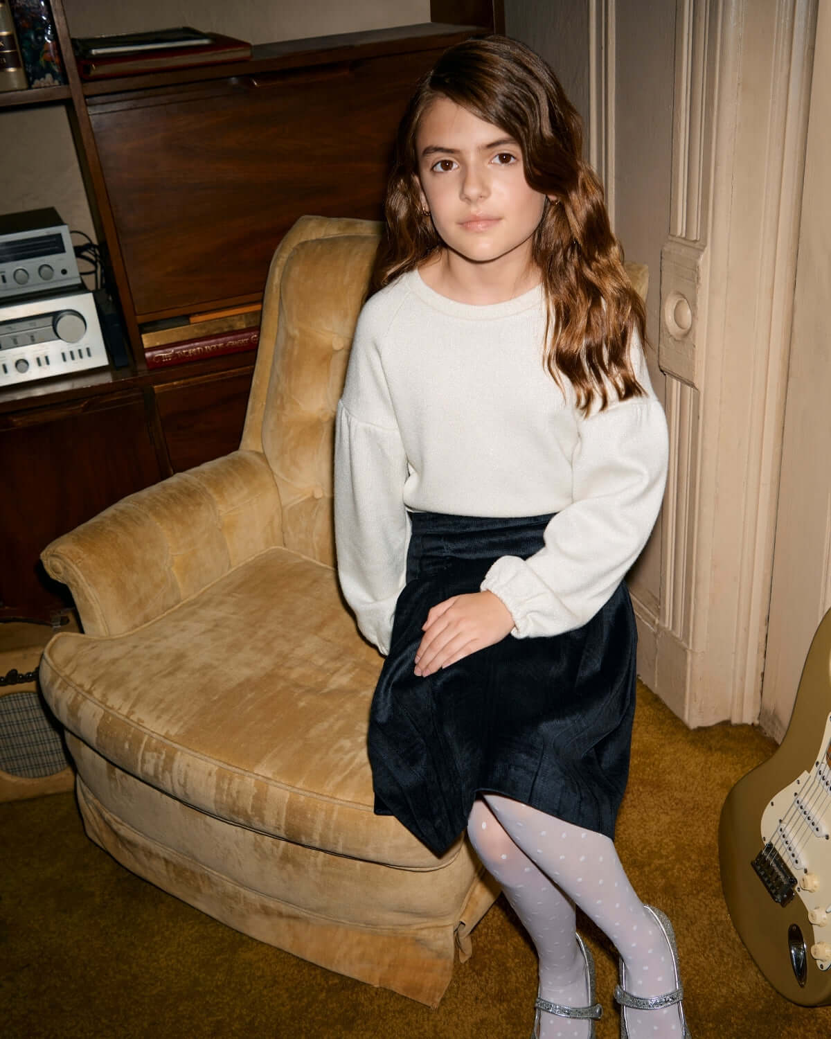 Child wearing Midnight Velvet Plaid Pleated Skirt with a cozy sweater, seated on a vintage chair.