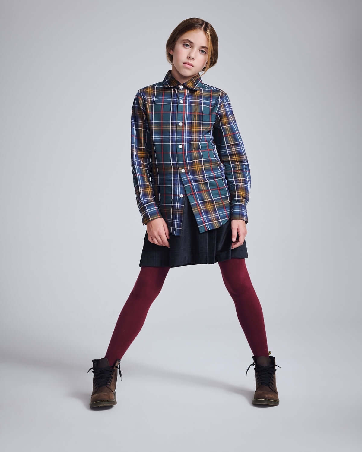 Girl wearing a plaid shirt with a Midnight Velvet Plaid Pleated Skirt and maroon tights, posing confidently.