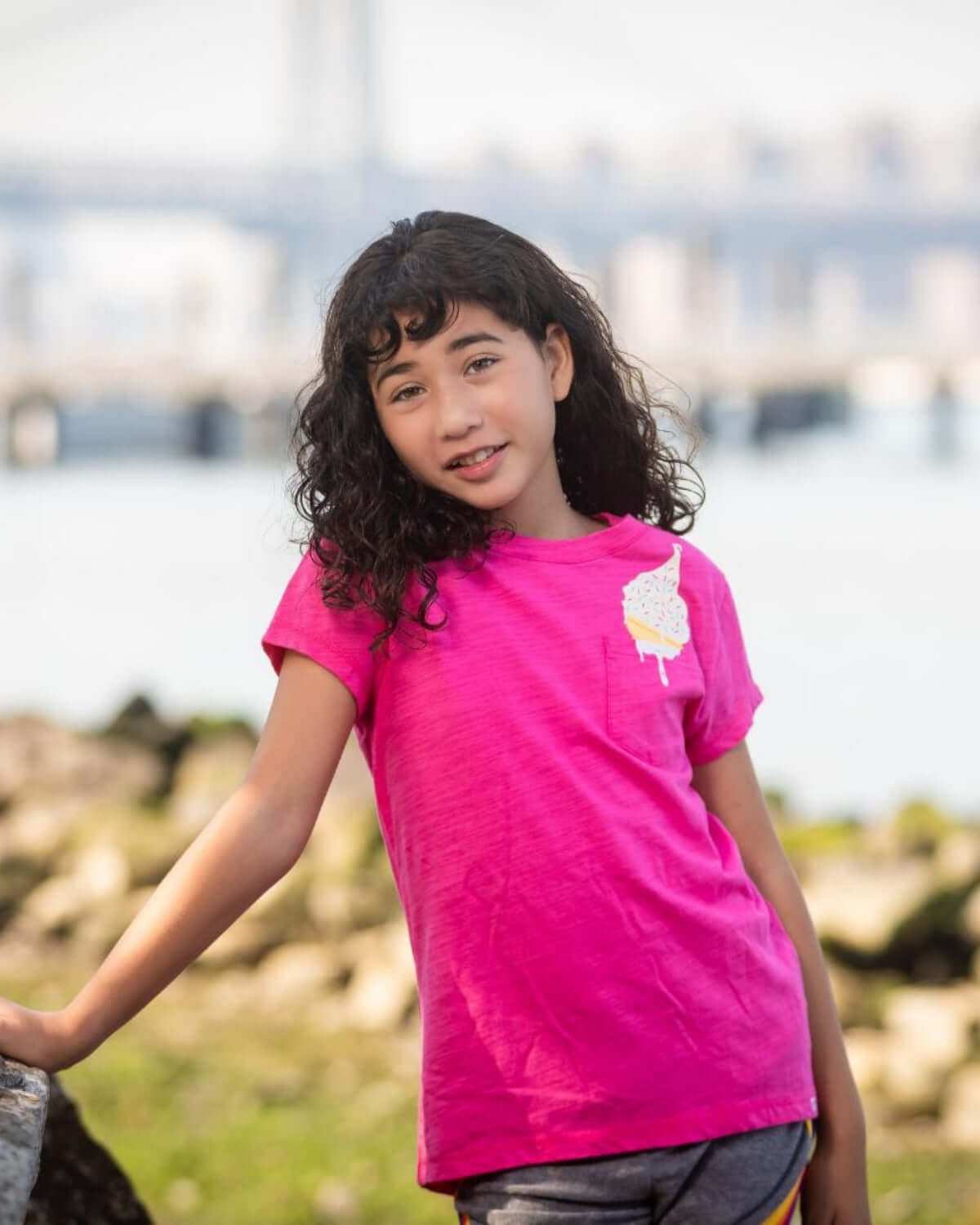Girl in vibrant fuchsia Pocket Ice Cream Pocket Tee smiling outdoors near water.