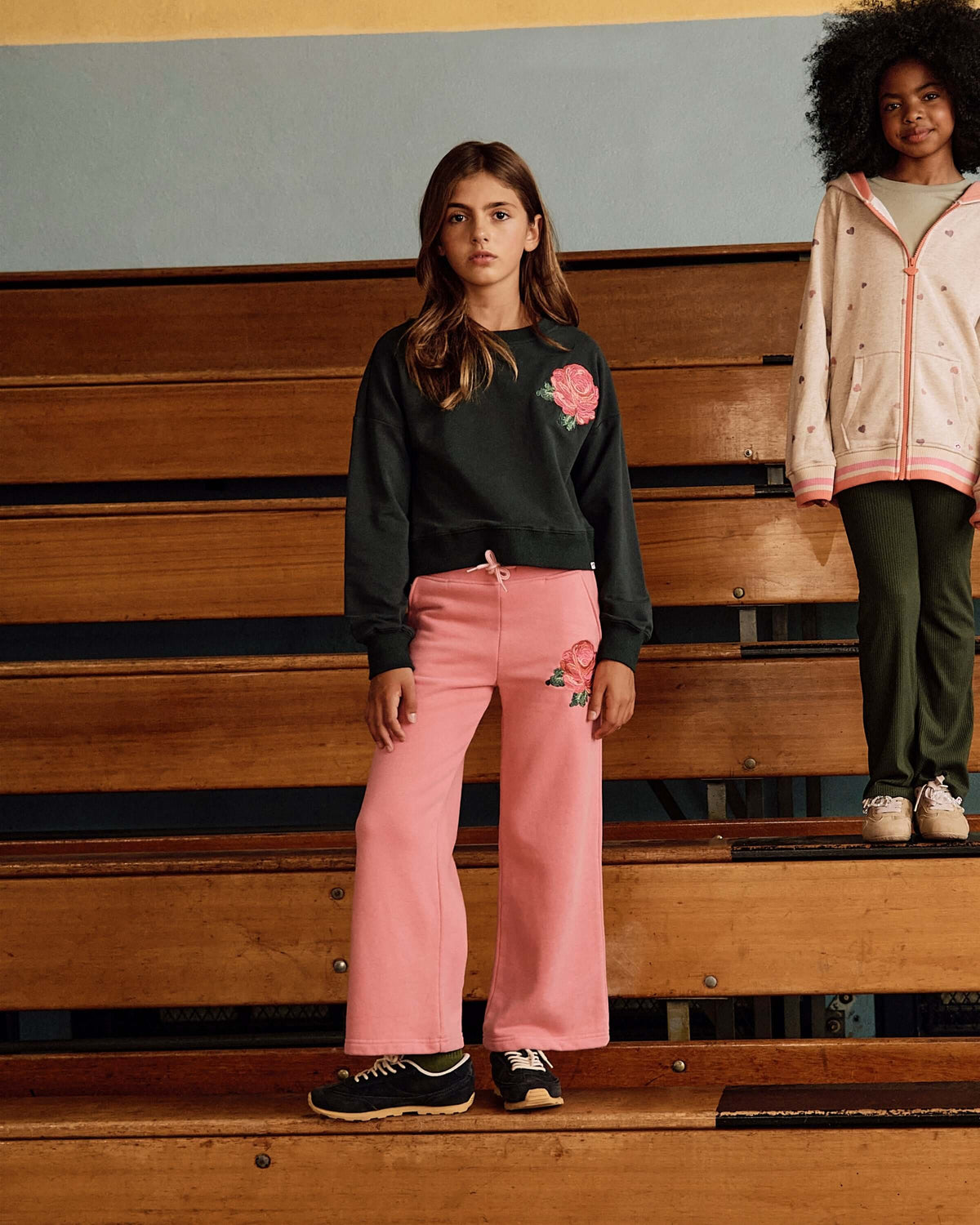 Rose Samantha Sweatpants in pink worn by a girl on bleachers, paired with a black sweatshirt and stylish sneakers.