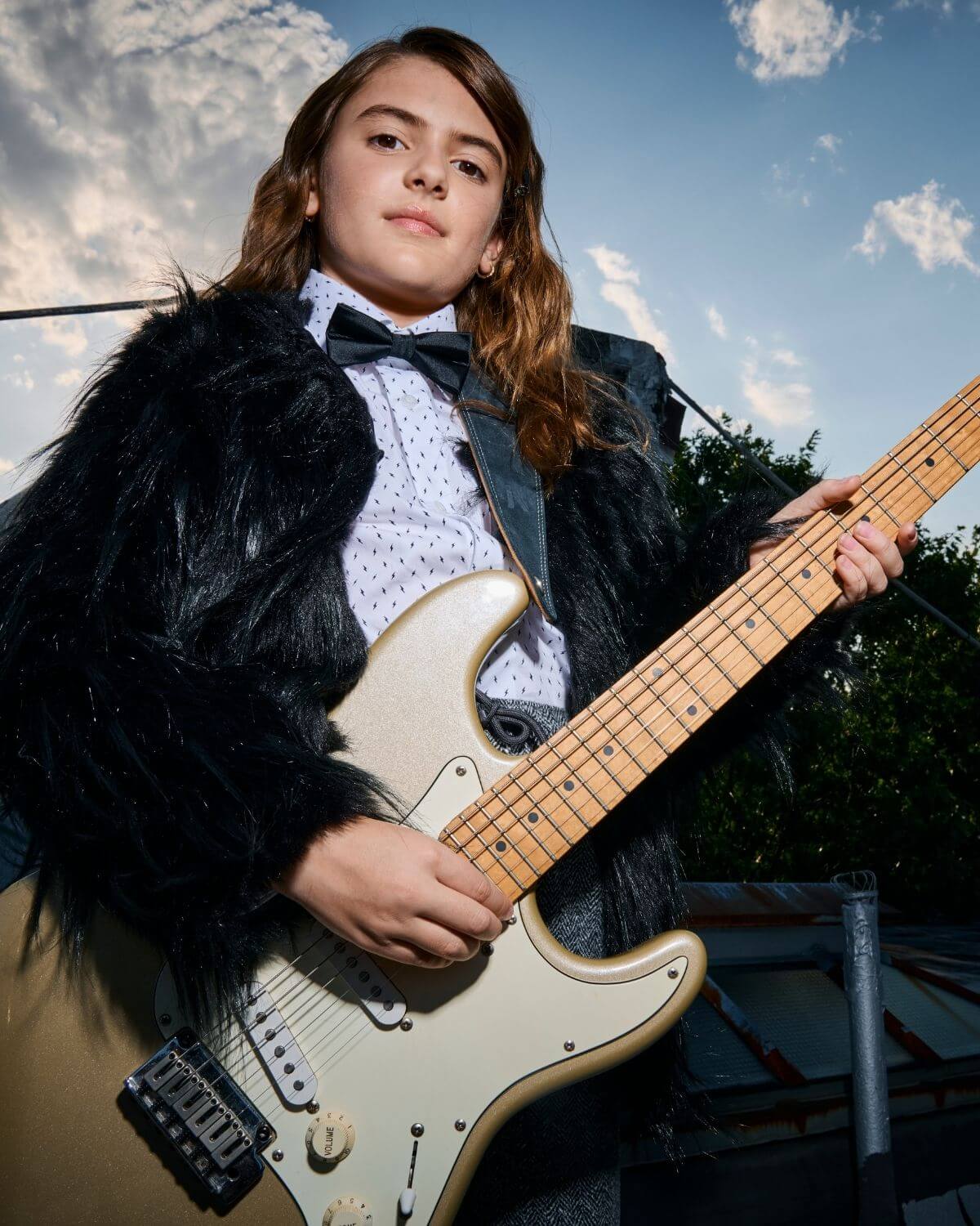 Child wearing a black silk bow tie while playing an electric guitar outdoors.