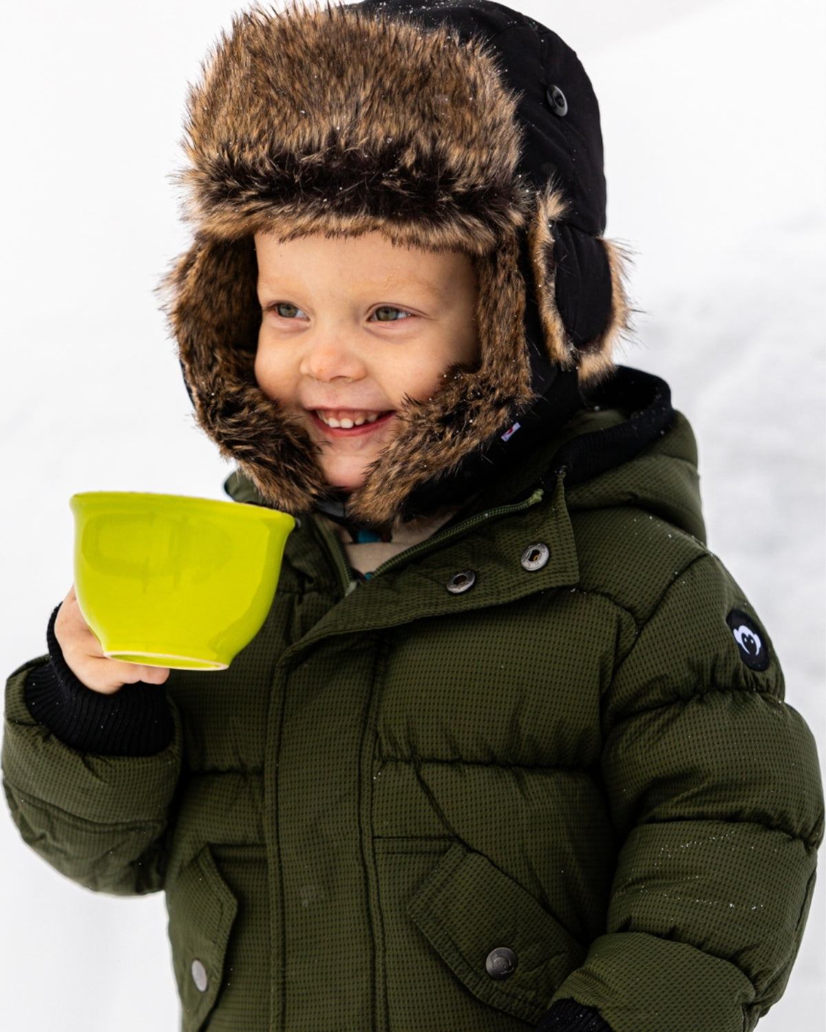 Cheerful kid in a faux fur-lined winter hat sipping from a bright bowl, ready to conquer winter adventures in stylish kids clothes.