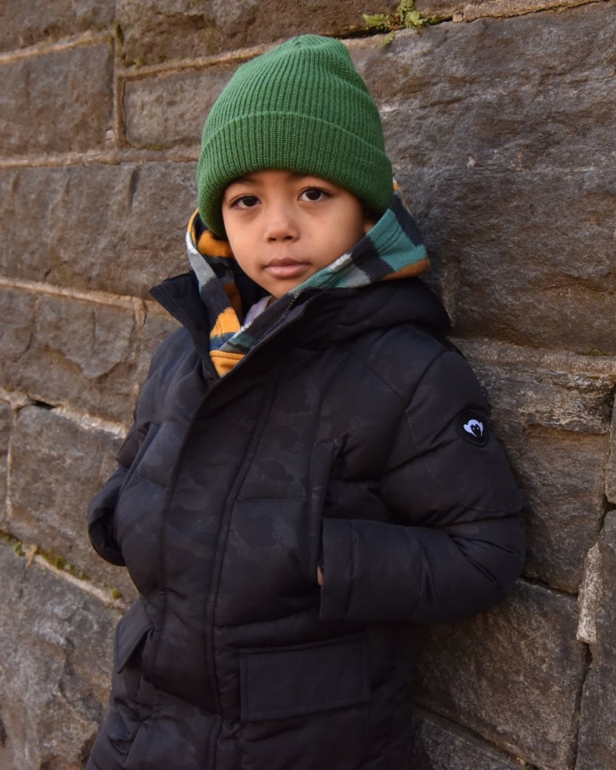 Trendy kid in a cozy black puffer coat and vibrant hunter green beanie, striking a confident pose against a stone wall.