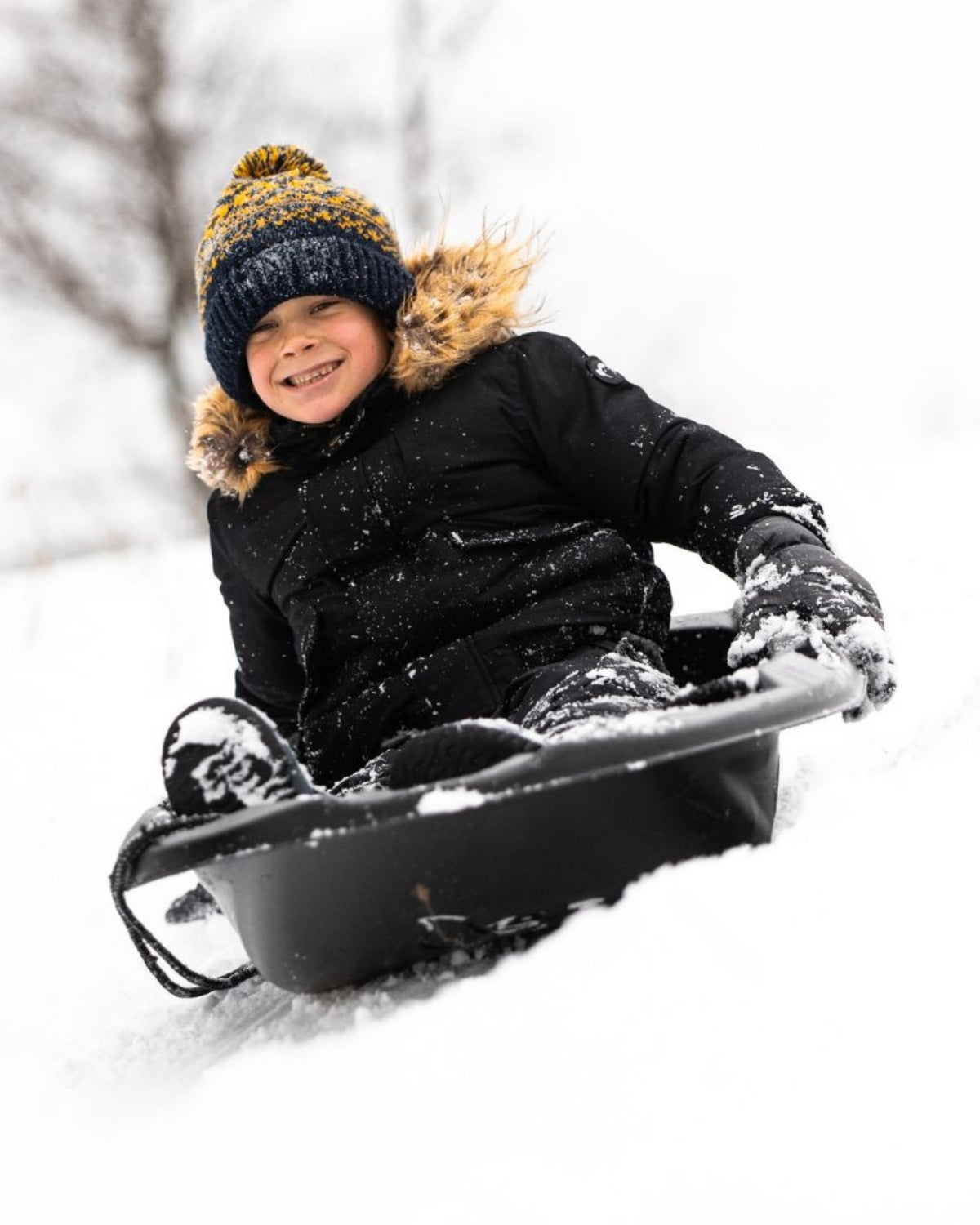A smiling boy in a cozy navy blue bush hat sledding through snowy fun, showcasing stylish kids clothes for winter adventures.