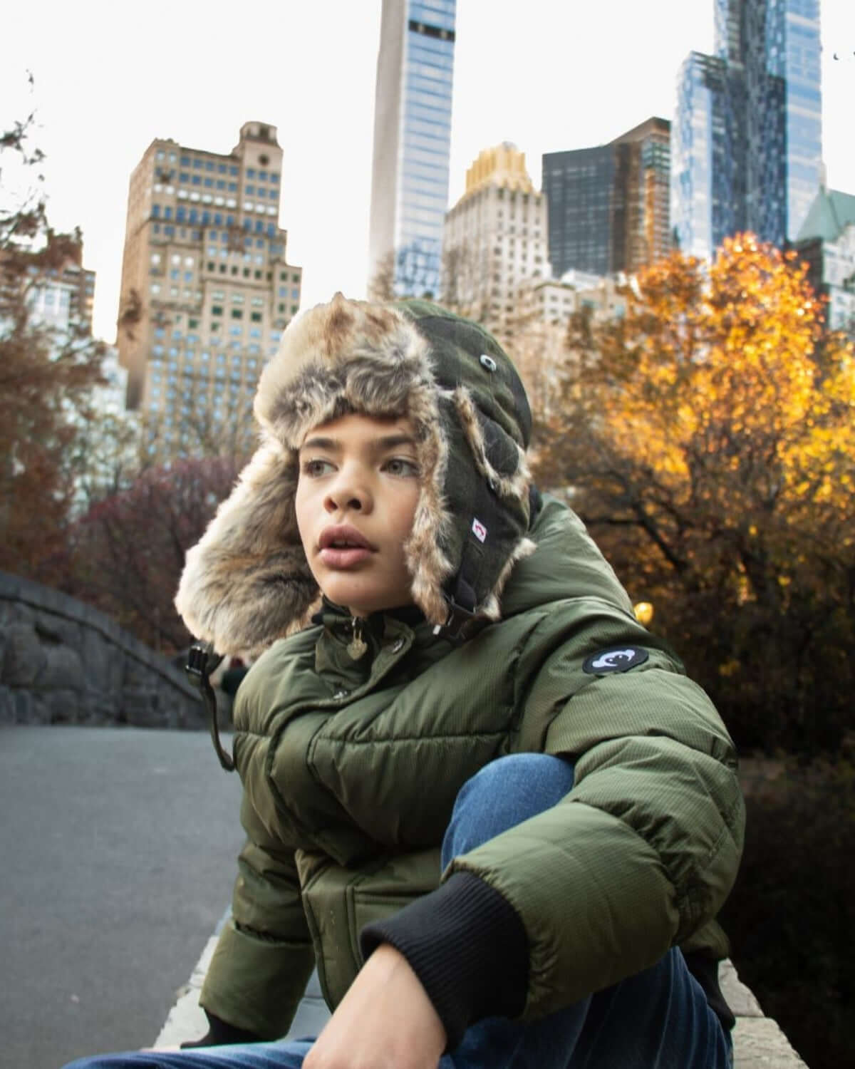 Stylish kid in green puffer jacket and trapper hat against a city skyline, showcasing trendy, warm kids clothes for winter.