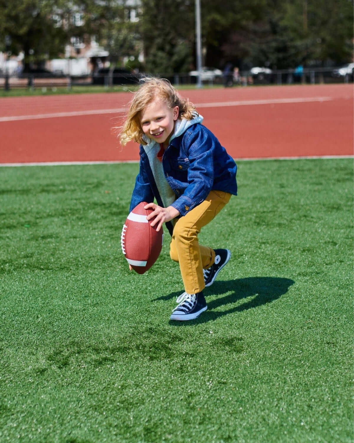A cheerful child in stylish British khaki pants, sprinting with a football on a sunny field, showcasing kids' activewear fun!
