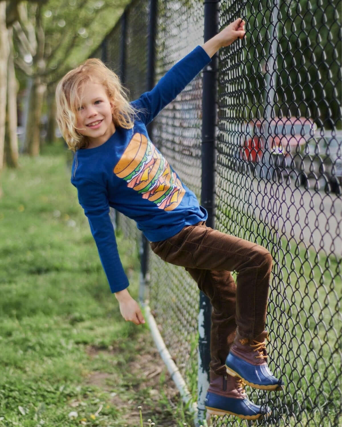 Playful boy in a blue burger tee and brown skinny cords, scaling a fence, ready for fun and style! Kids clothes from Appaman.