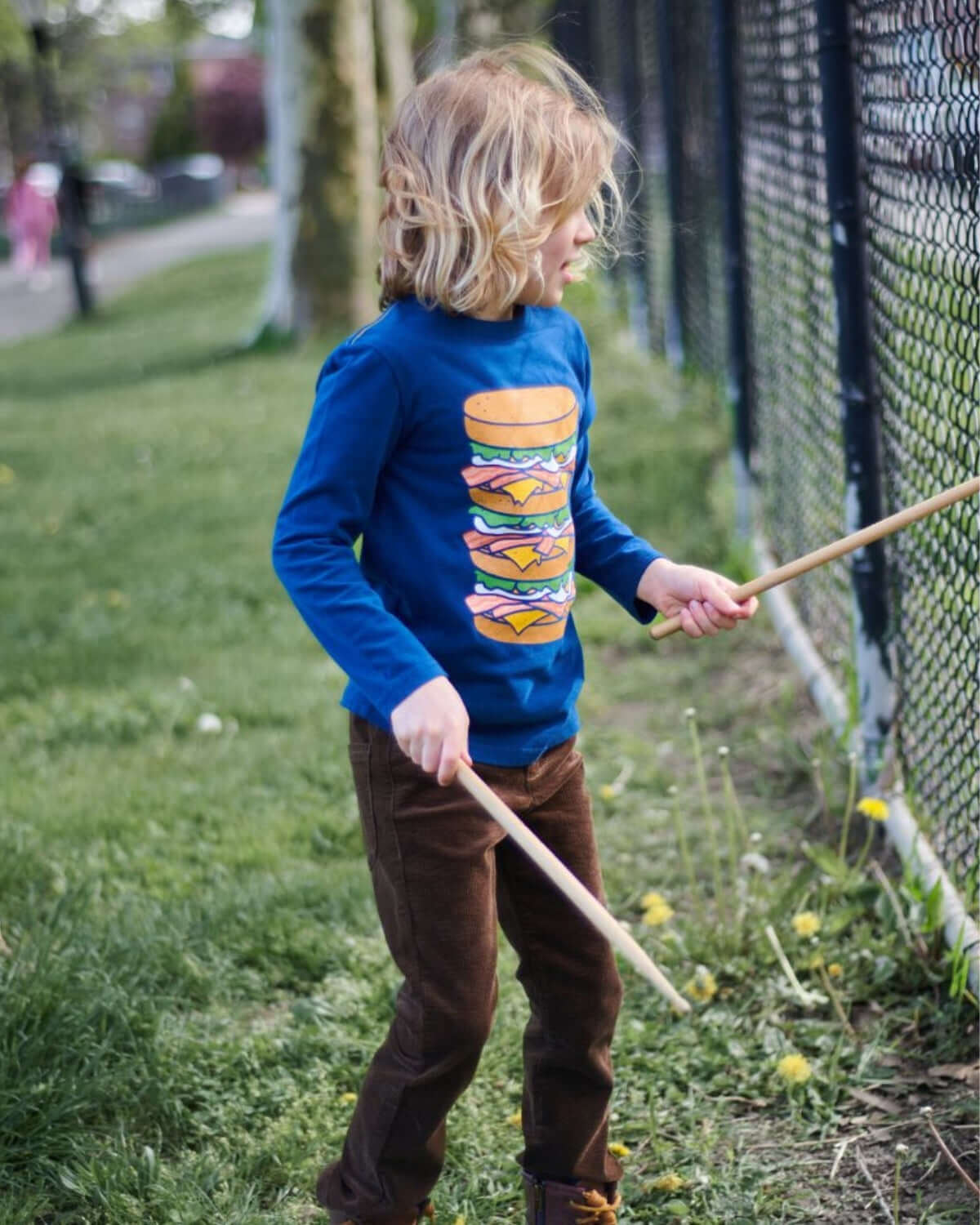 Stylish kid in a fun blue burger graphic tee and brown skinny cords, drumming with sticks in a sunny park.