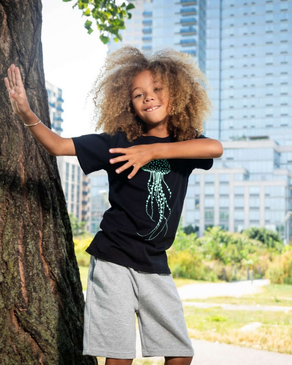 A playful child showcasing stylish boys clothes, wearing a black jellyfish tee and heather mist Preston shorts, outdoors in the city.