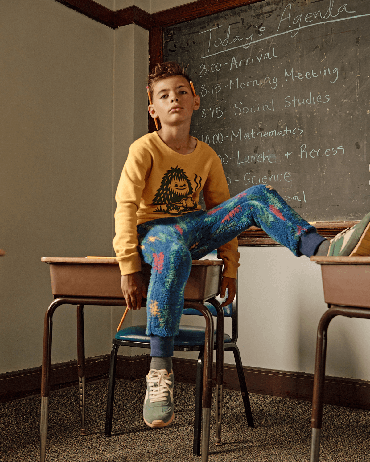 A stylish boy in a playful sweatshirt and vibrant sweats, showcasing modern kids' fashion in a classroom setting.