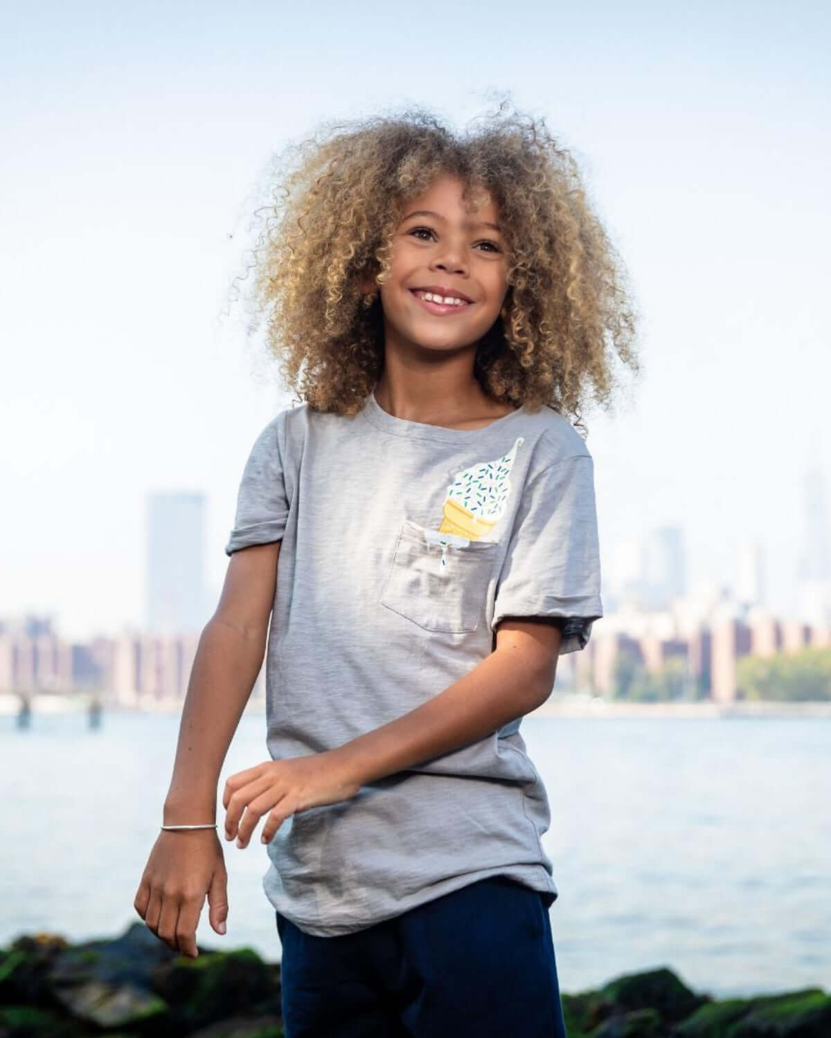 Joyful kid in a stylish gray tee with a pocket design, ready for summer fun against an urban backdrop.