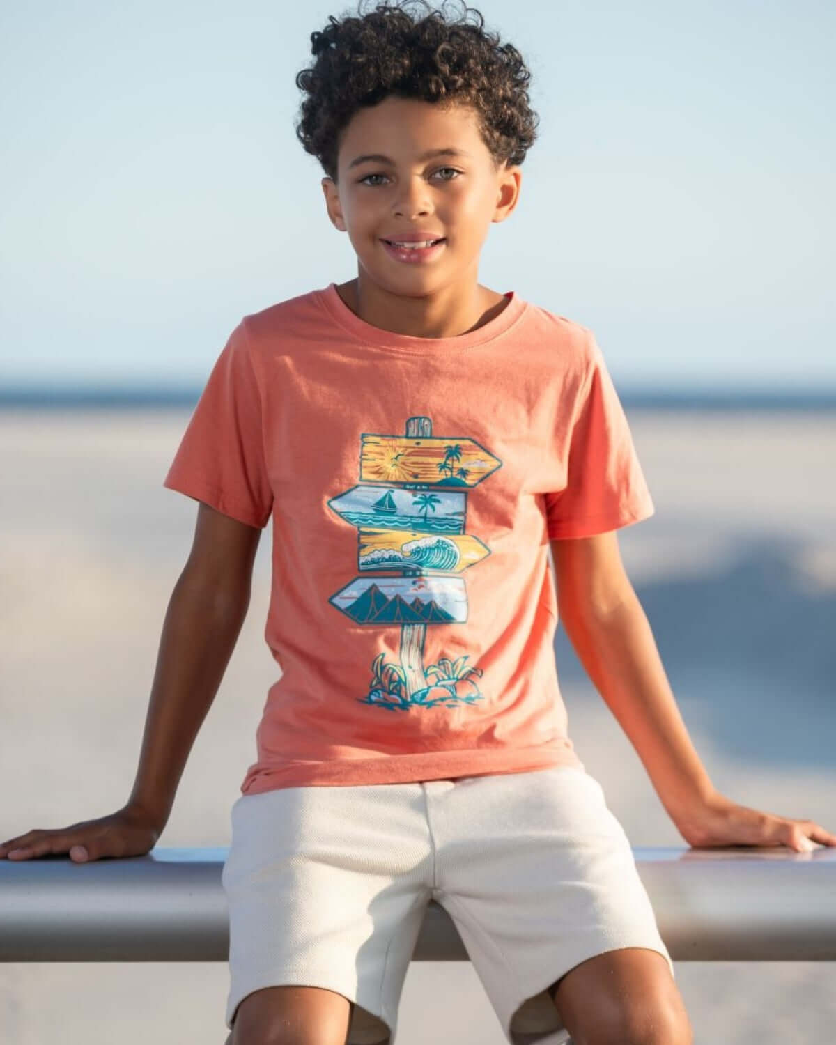 Boy wearing off white resort shorts and a coral t-shirt with a signpost graphic at the beach.