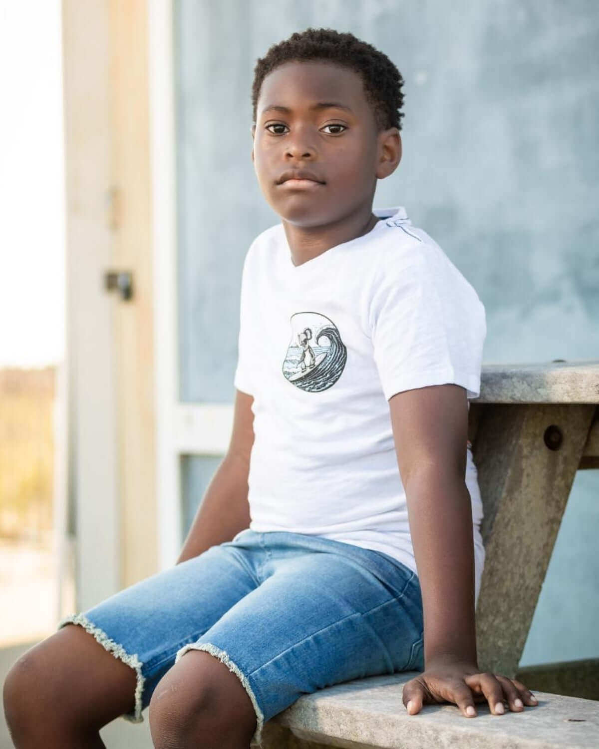 Boy wearing Sun Wash Denim Shorts and a white t-shirt, sitting on a bench outdoors.