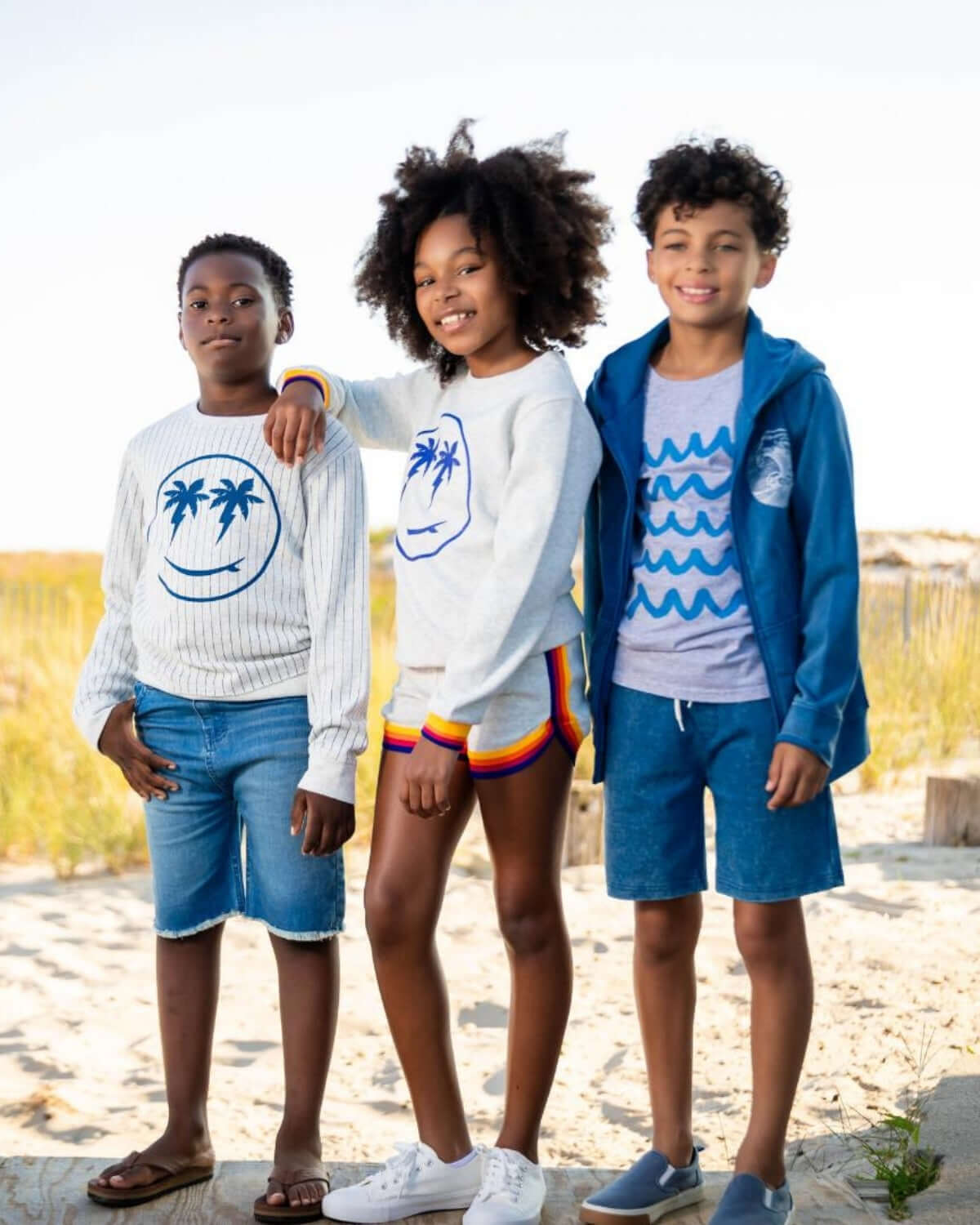 Three kids in stylish summer outfits, smiling on the beach, ready for sunny adventures in quality kids clothes.