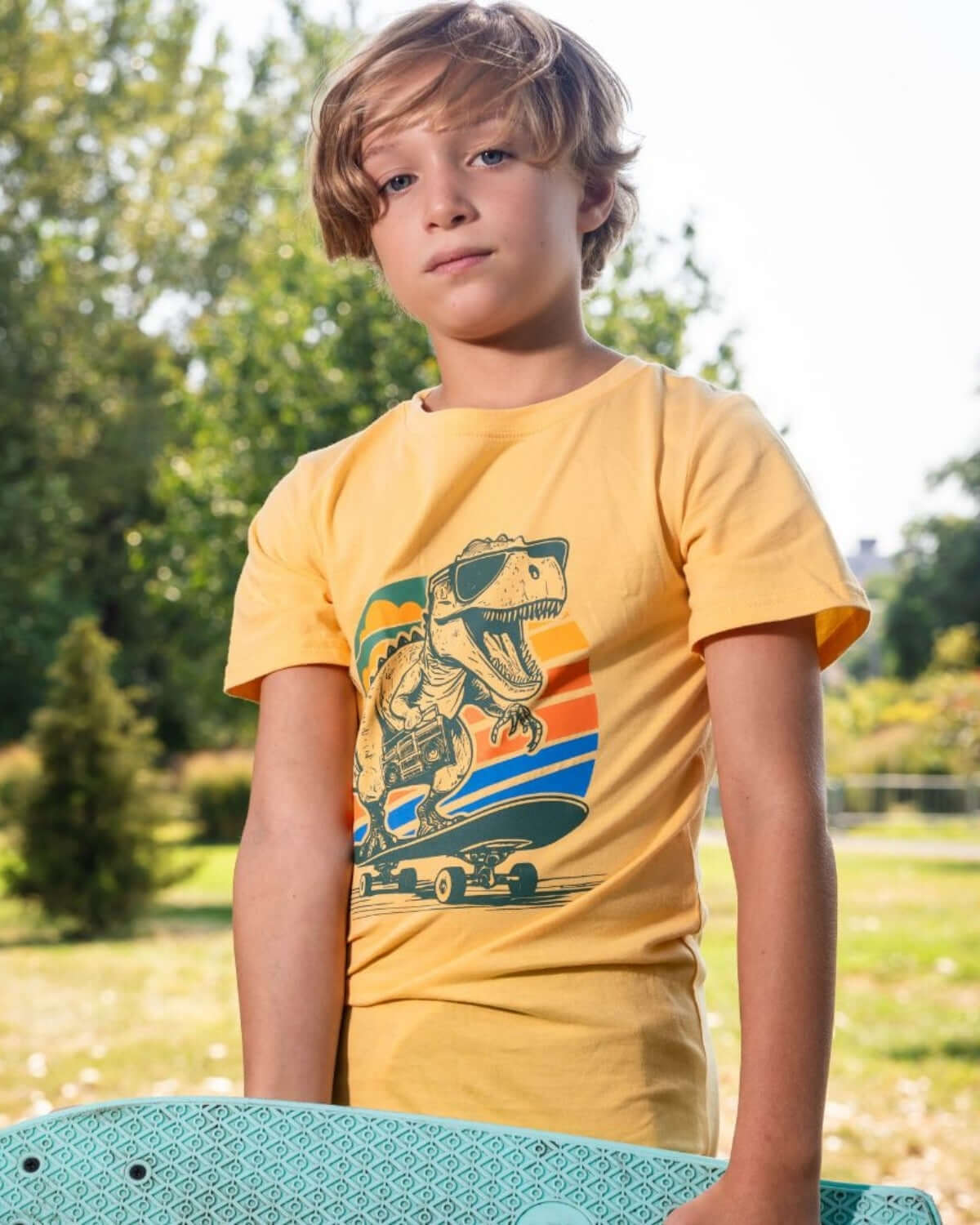 Stylish boy in Dino Rock graphic tee, ready for adventures with skate in park—fun kids clothes for back to school!