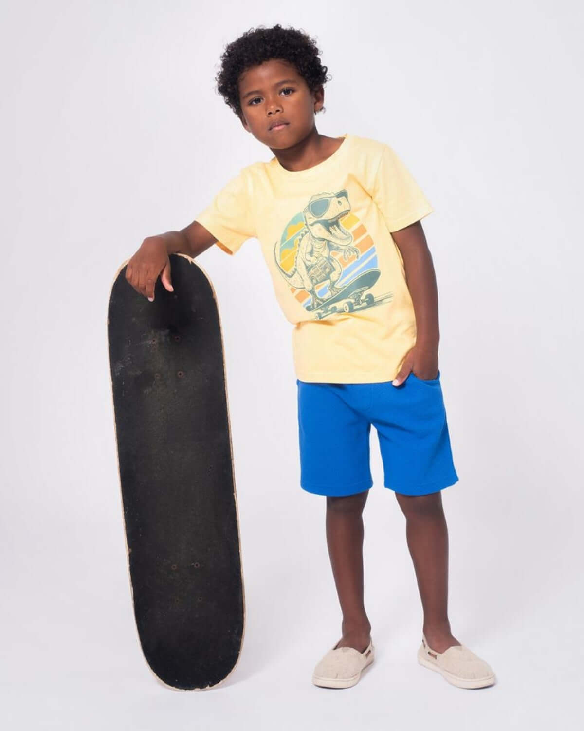 Stylish boy rocking a yellow Dino Rock tee with blue shorts and skateboard, showcasing cool kids clothes for fun adventures.