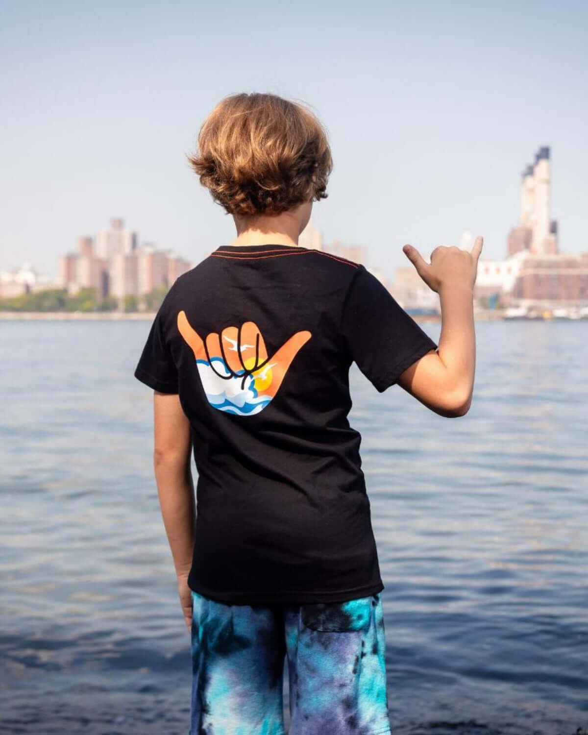 Boy wearing Hang Loose Graphic Tee with hang loose hand signal by the water, perfect for beach days.