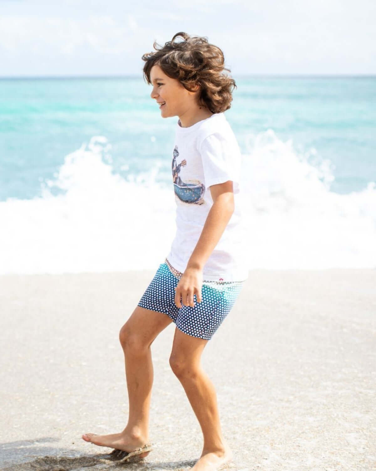 A happy boy splashes in ocean waves wearing the Madagas-Star Graphic Tee and stylish swim shorts, perfect for summer fun!