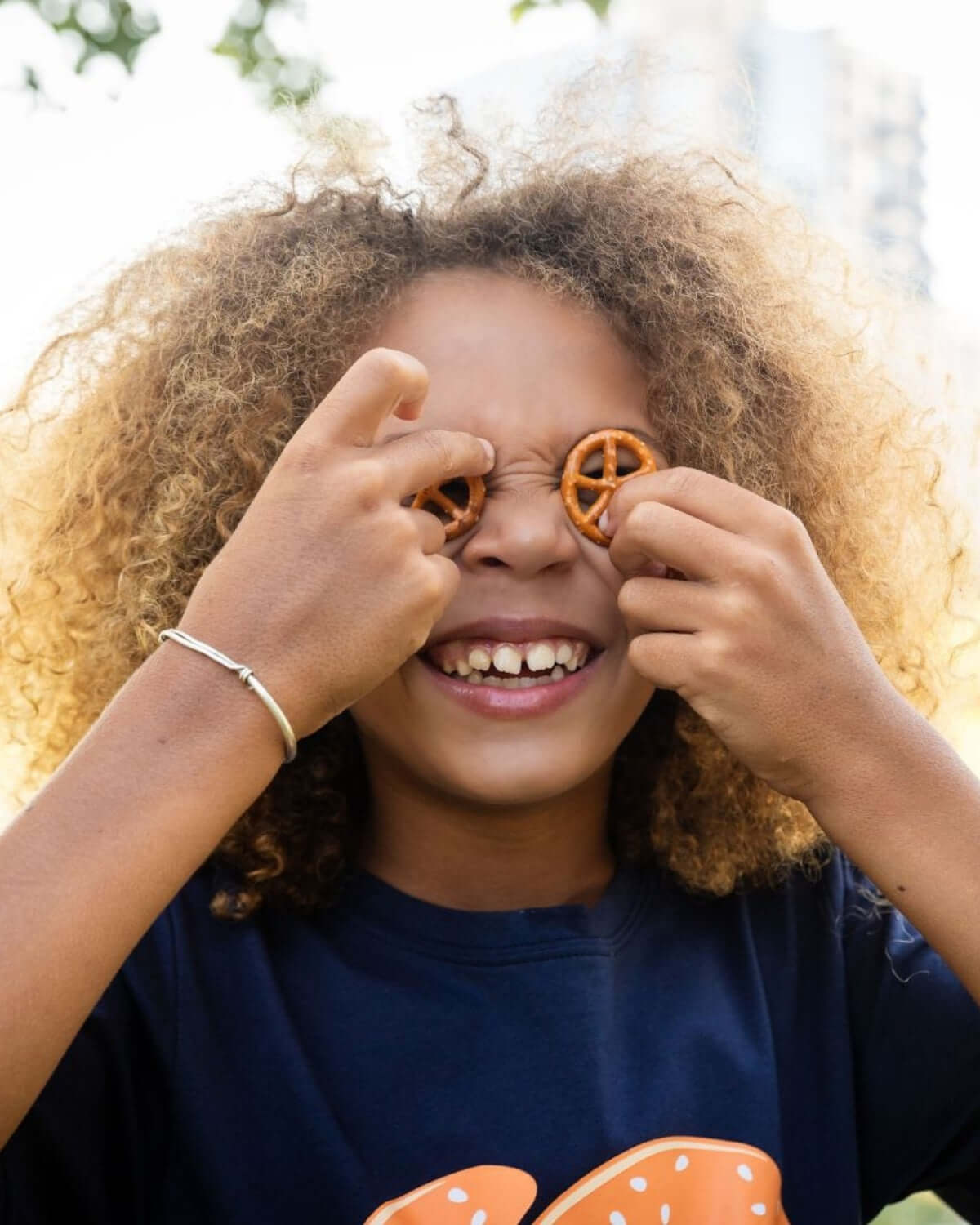 Joyful child wearing a pretzel graphic tee playfully holding pretzels in front of eyes, showcasing fun kids fashion.