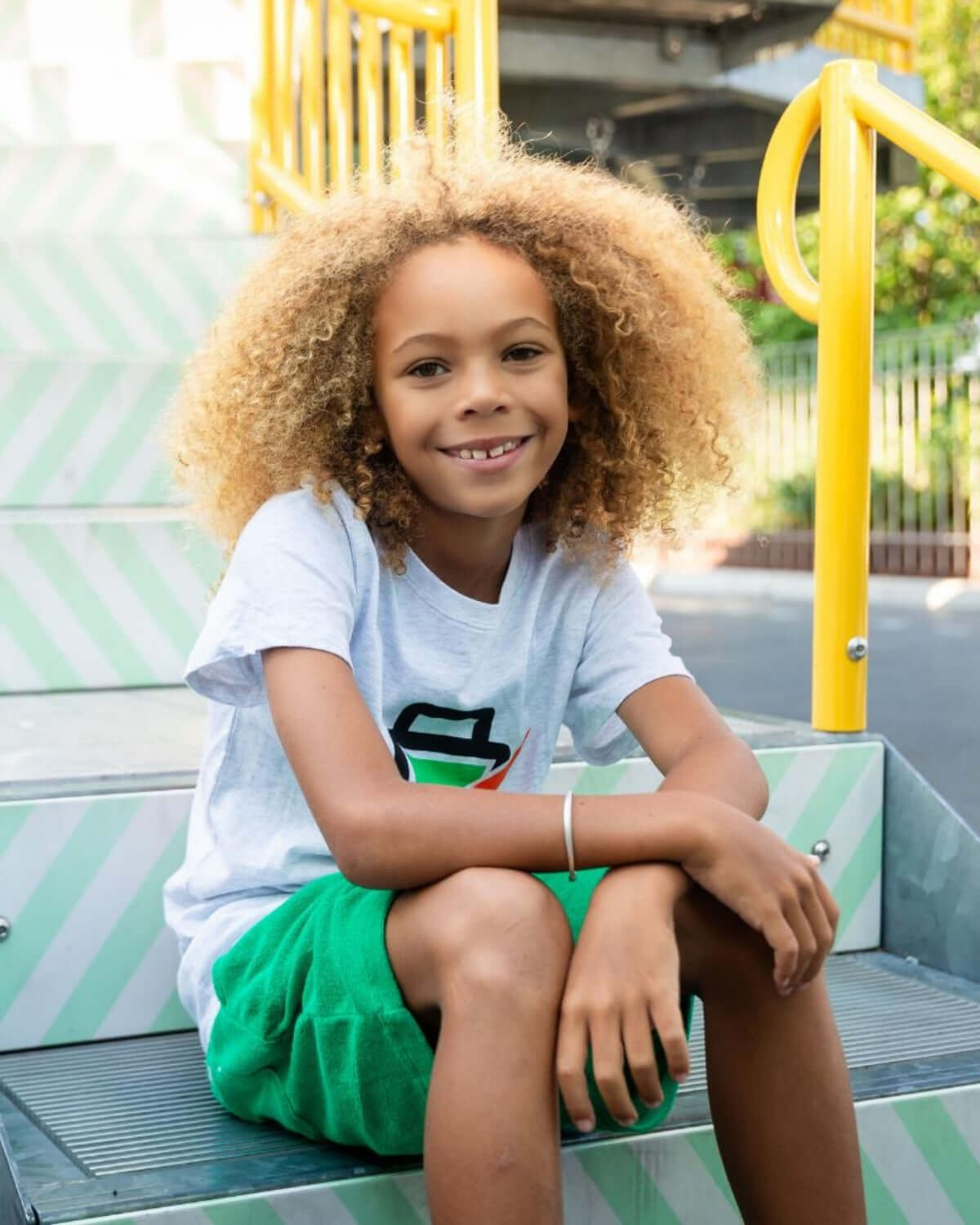 Cheerful child wearing a Recharged Graphic Tee with a friendly battery design, sitting on colorful stairs.