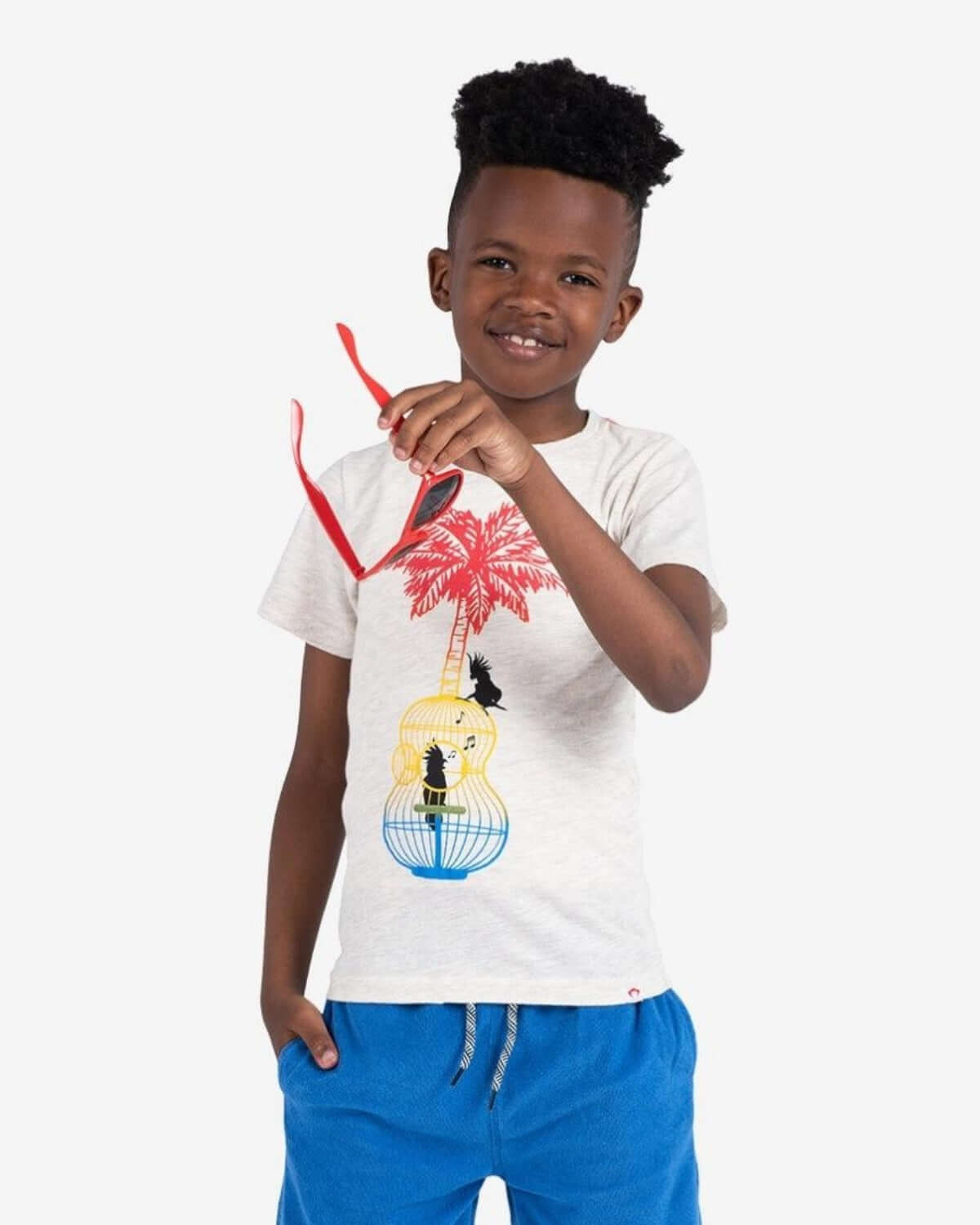 Boy wearing Song Birds Graphic Tee, holding red sunglasses with palm and guitar illustration.