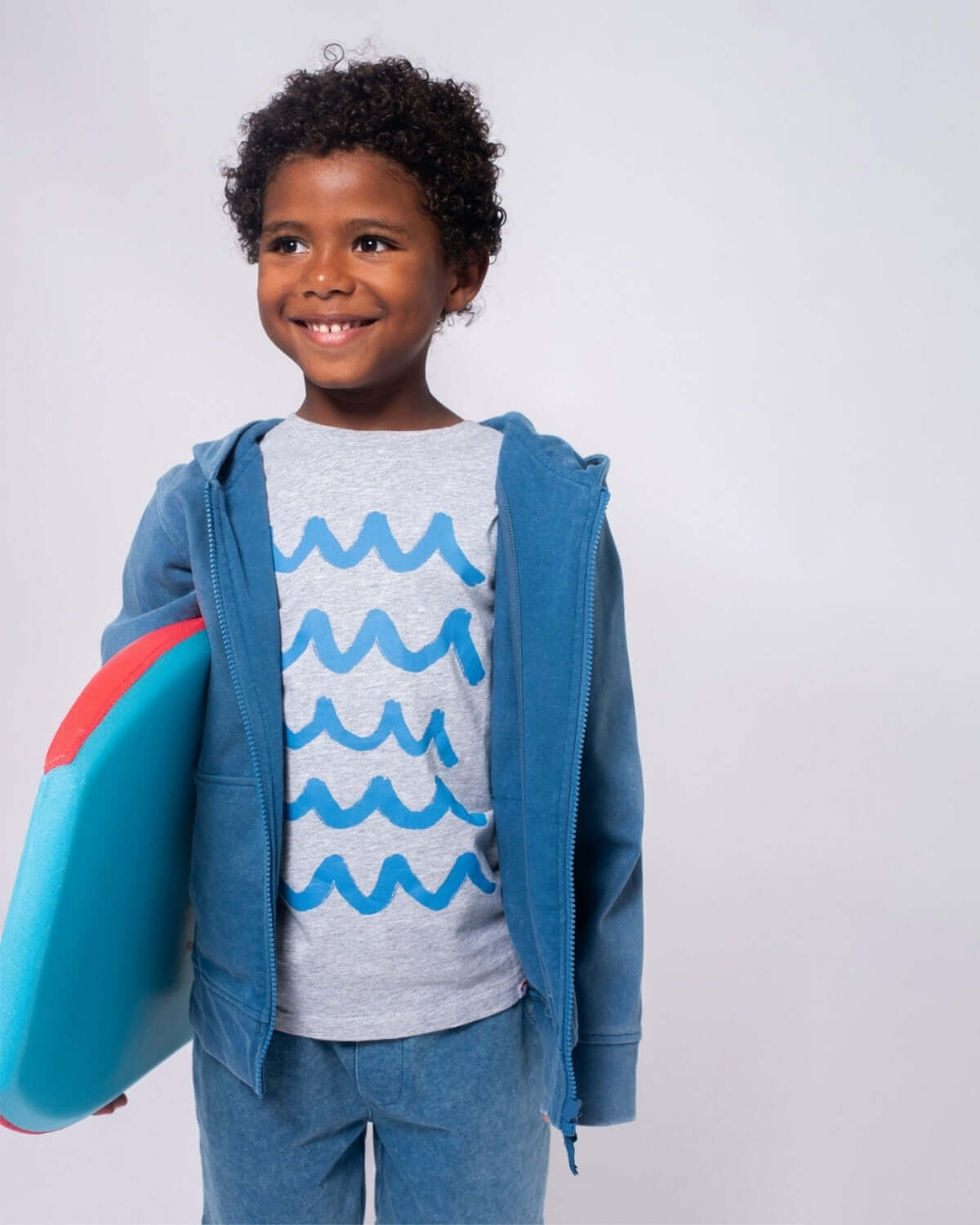 Smiling boy wearing a Waves Graphic Tee with ocean wave design, holding a foam surfboard, ready for beach fun.